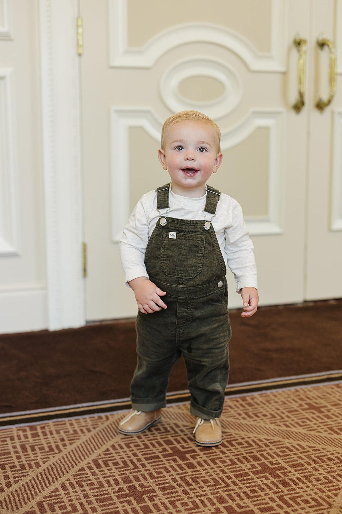 A toddler wearing a white long-sleeve shirt and olive green overalls stands on a patterned carpet.