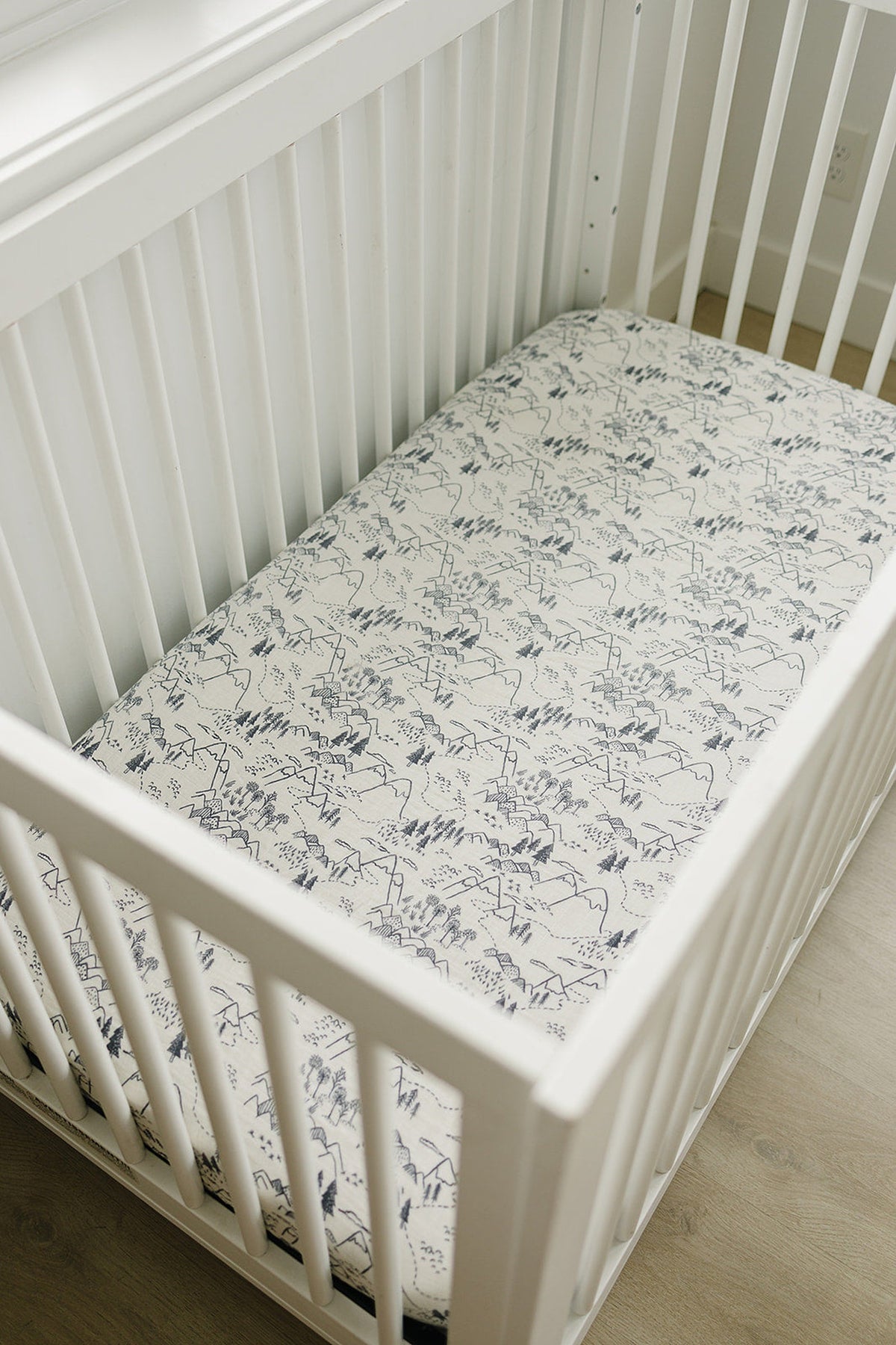 A muslin crib sheet featuring a black and white mountain and forest design fits snugly in a white wooden crib.