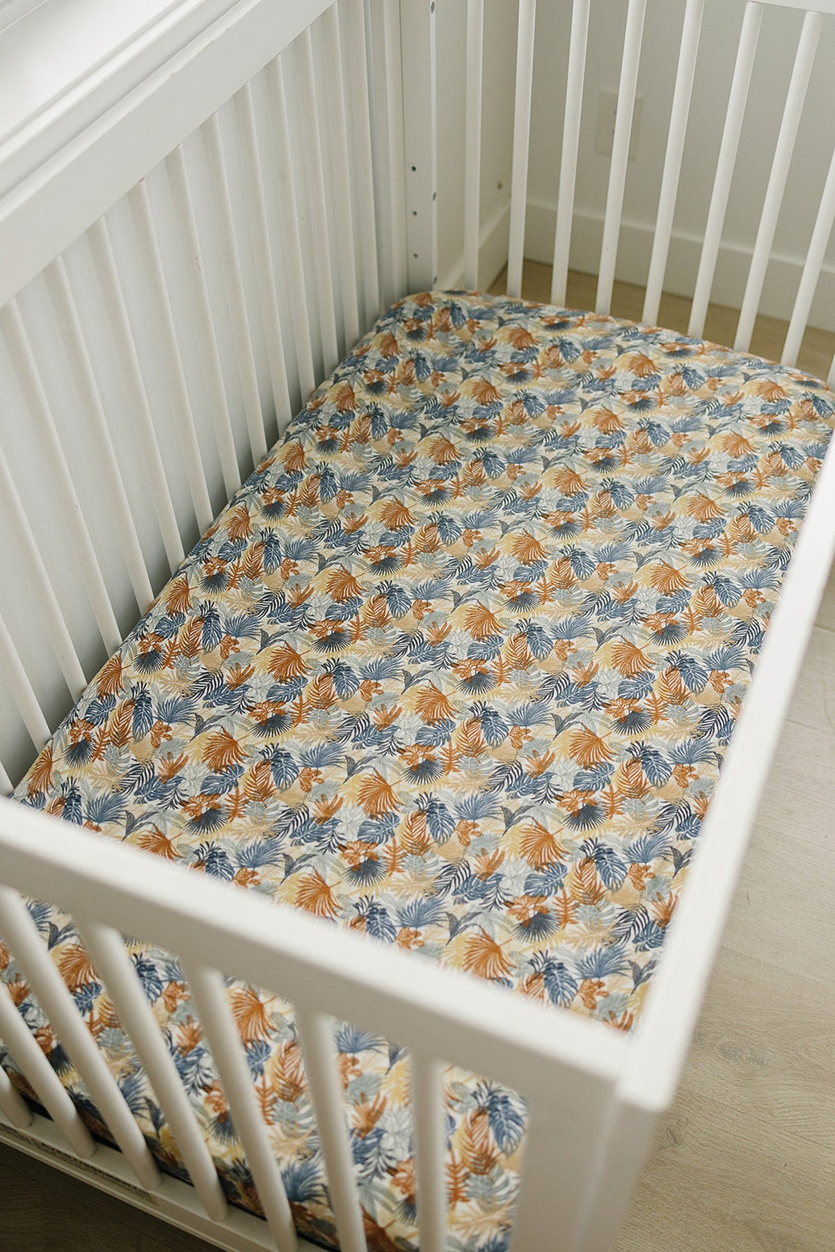 White crib with a colorful palm leaf patterned muslin sheet in orange, blue, and beige hues.
