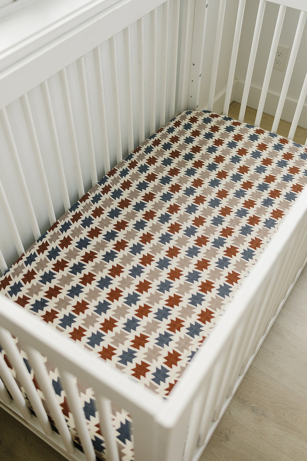 A white crib features a colorful Western Aztec muslin crib sheet with geometric patterns in red, blue,.