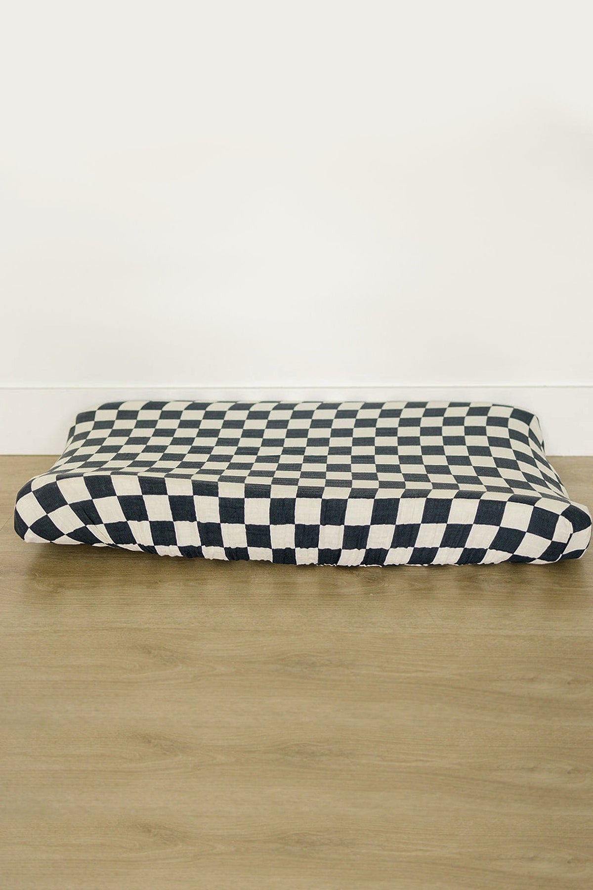 Charcoal checkered muslin changing pad cover on wooden floor, featuring a modern design with elasticized edges for a snug fit.
