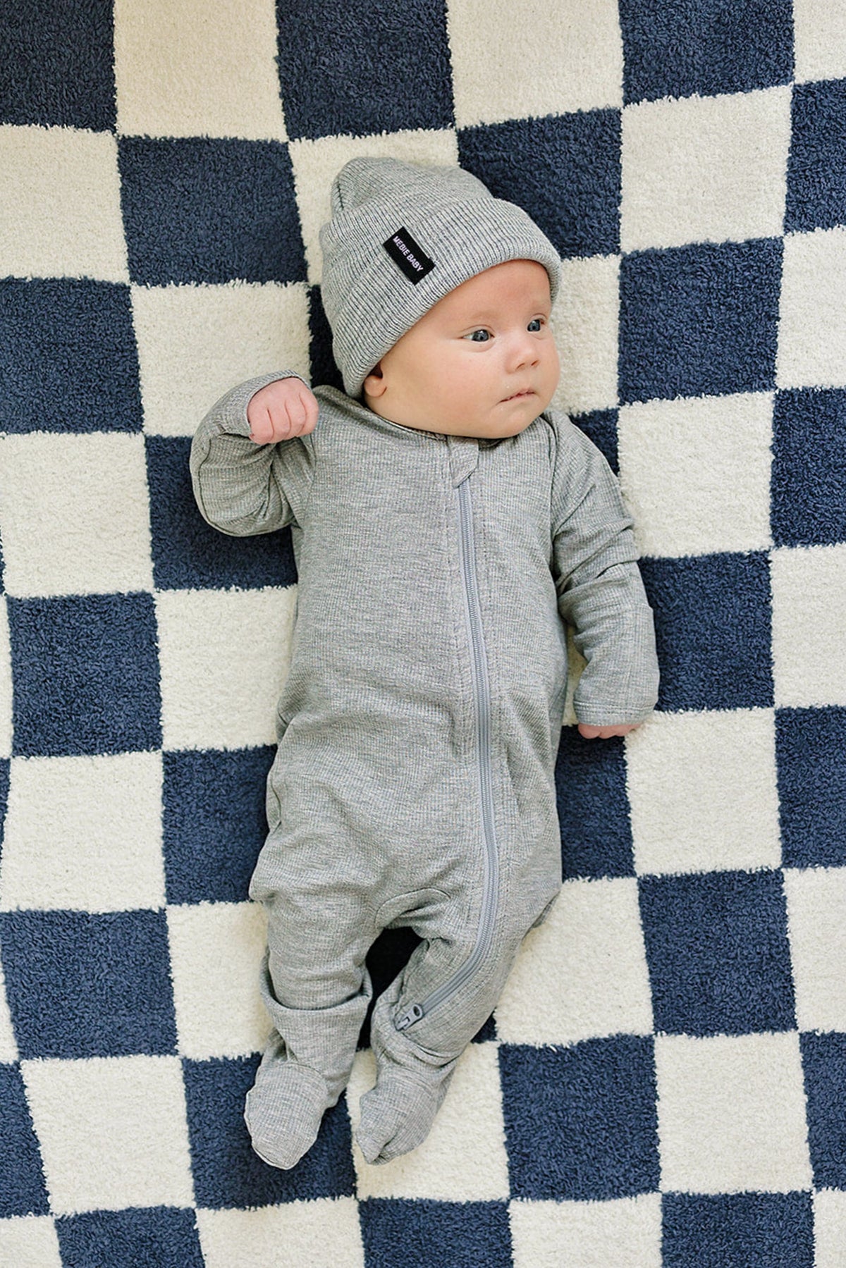 A baby in a grey ribbed zipper pajama and matching hat lies on a blue-and-white checkered blanket.