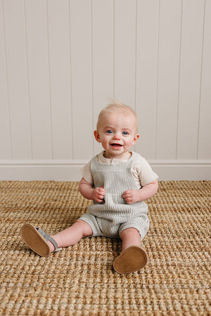 A smiling baby in heather grey knit overalls sits on a textured rug against a light-paneled wall.