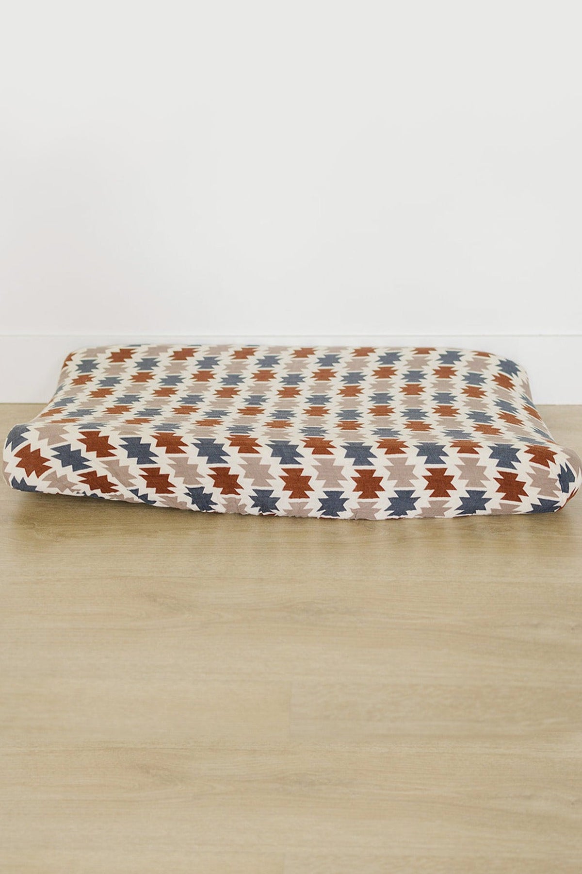 Western Aztec muslin changing pad cover with a geometric pattern in blue, rust, and beige on a wooden floor.