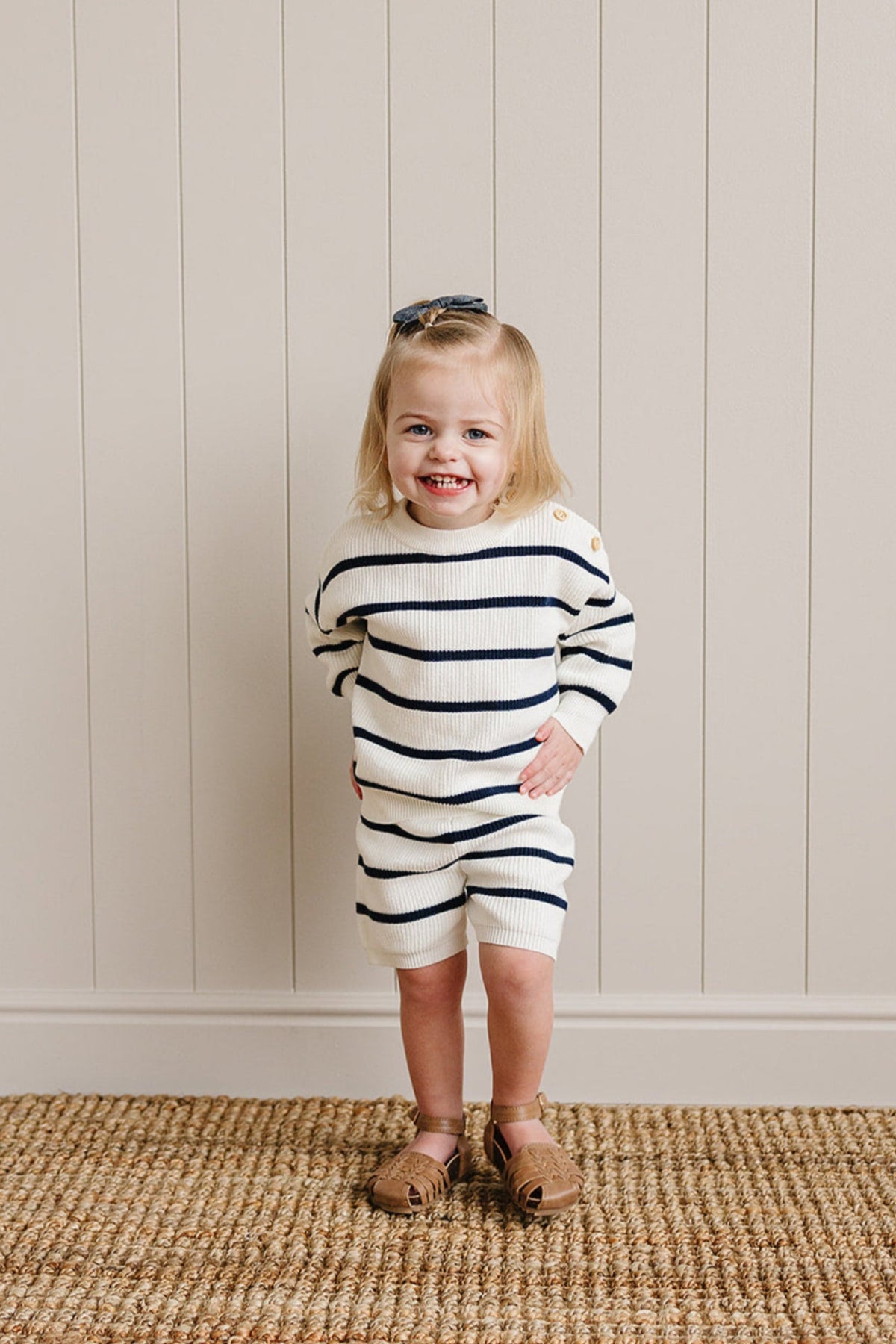 Toddler wearing a navy and white striped knit sweater and shorts, smiling, standing on a woven rug.
