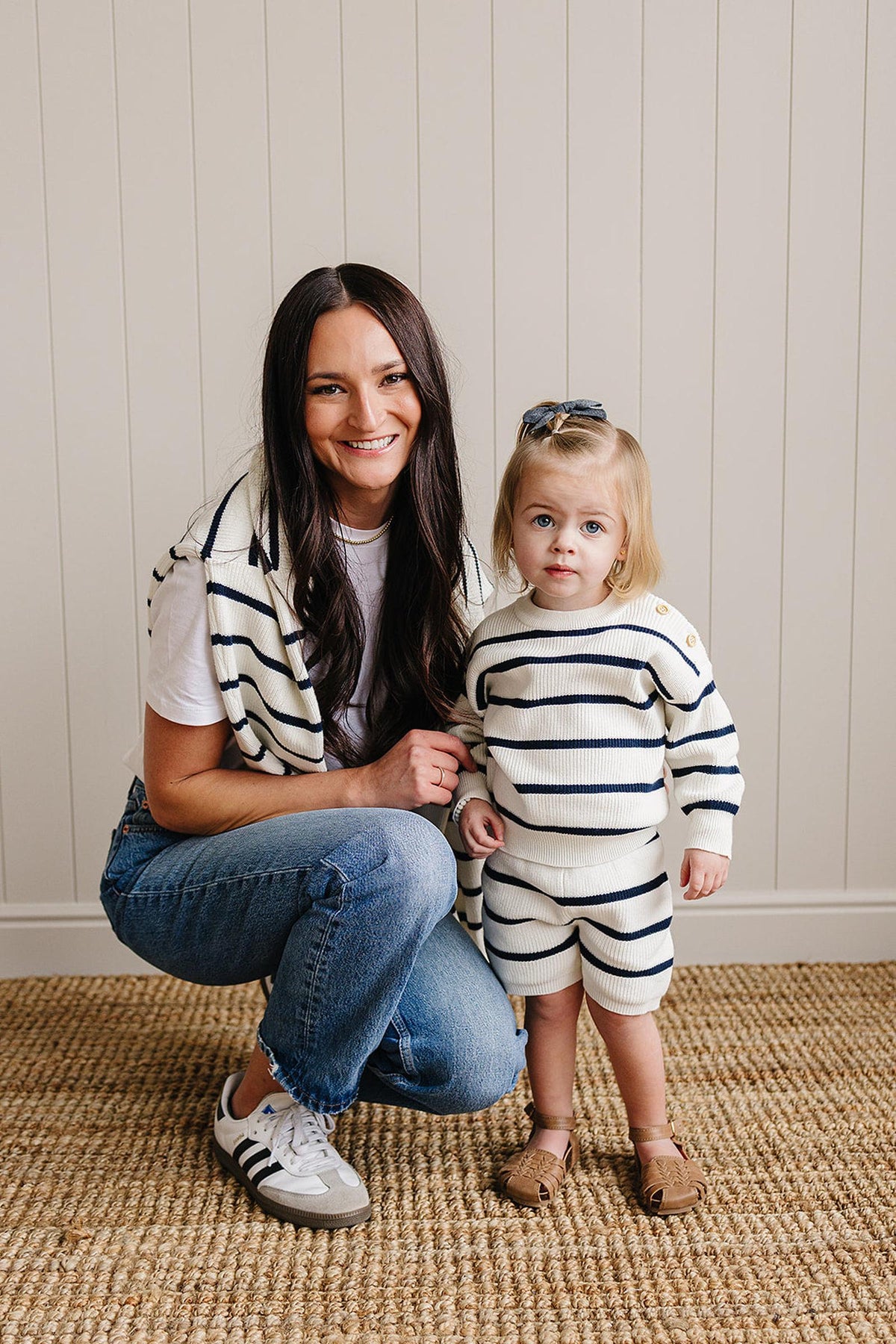 A woman and child wear matching navy and white striped knit outfits, smiling in a cozy, neutral room.