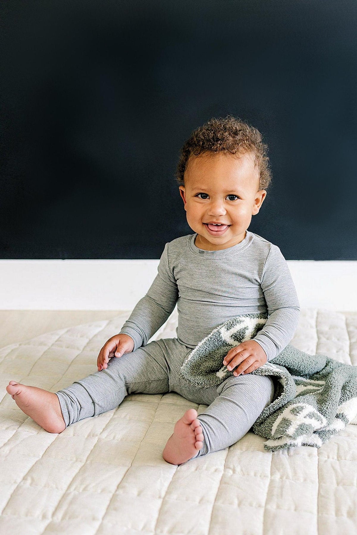 A smiling toddler in a grey ribbed pajama set sits on a quilted surface, holding a soft blanket.