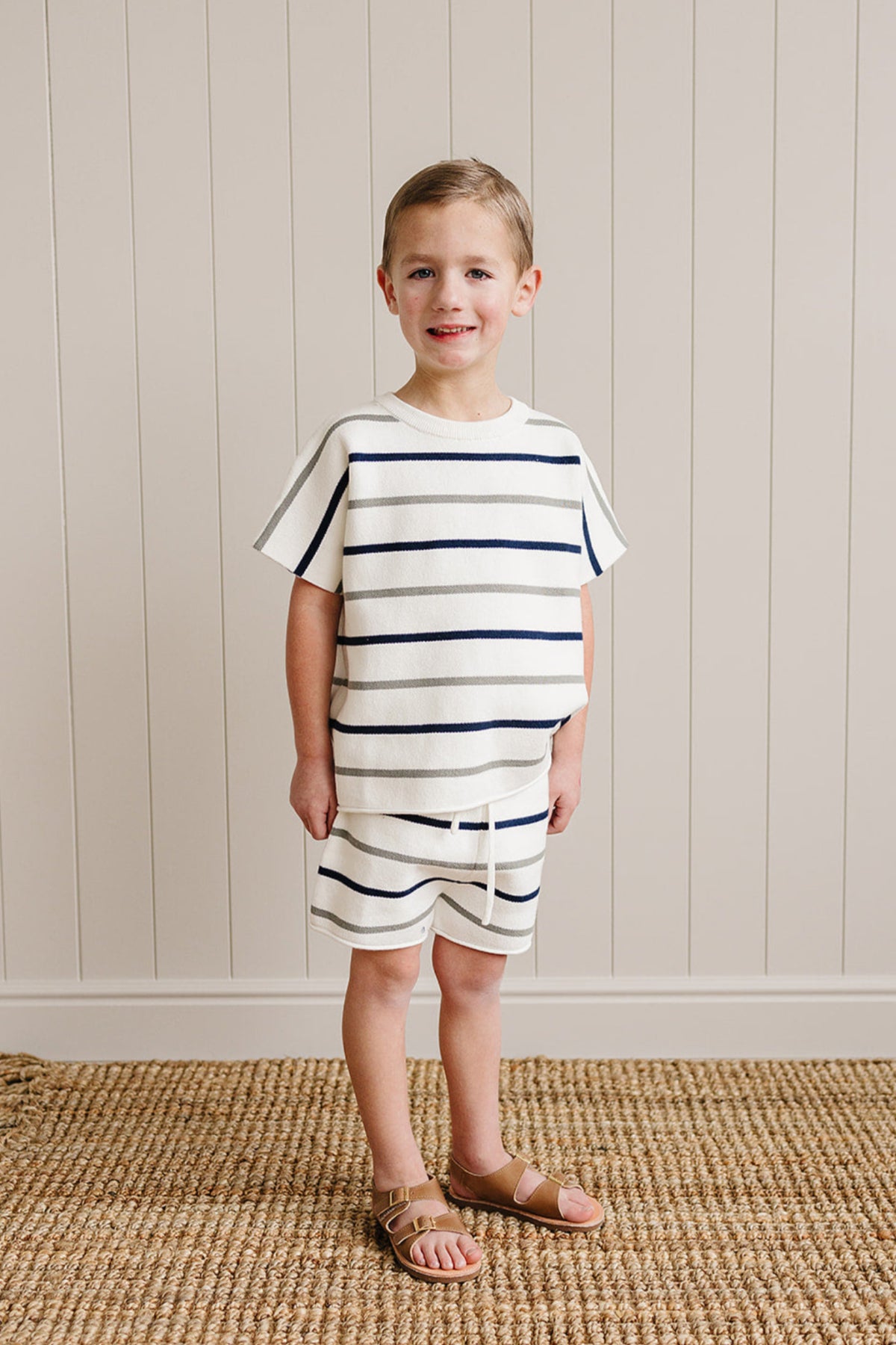 A child wearing a white knit outfit with navy and gray stripes stands on a woven rug.