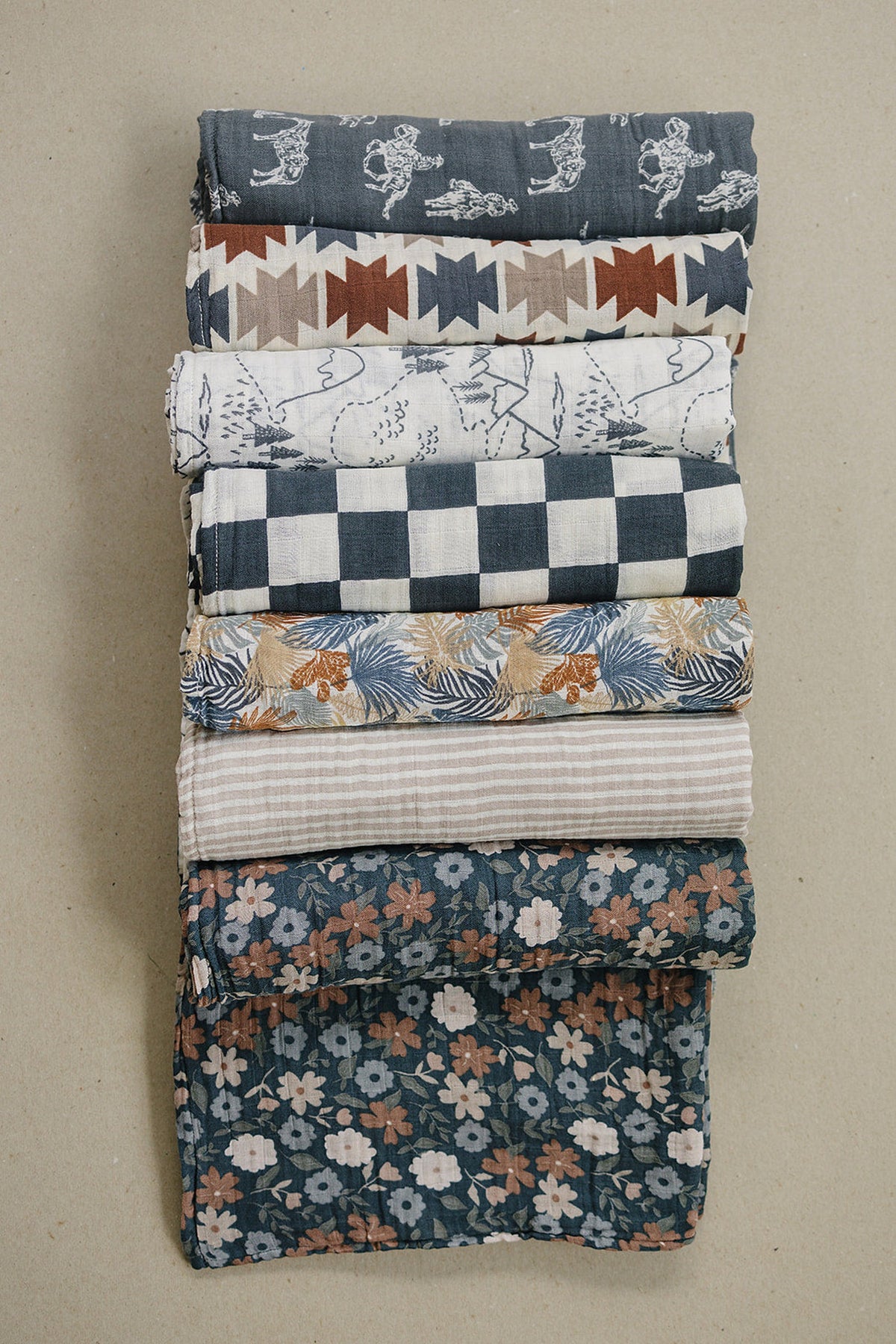 A stack of muslin swaddle blankets in various patterns, including floral, striped, and geometric designs.