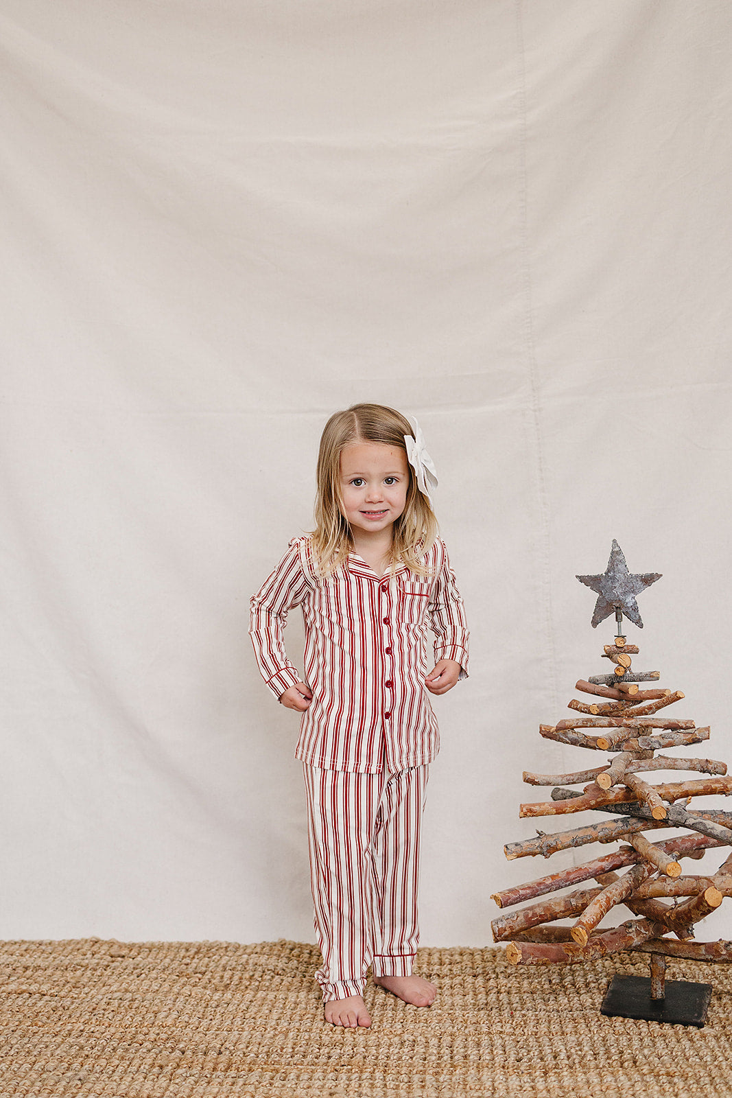 A child stands smiling in a red and white striped collared pajama set, made of soft fabric, next to a rustic wooden Christmas tree.