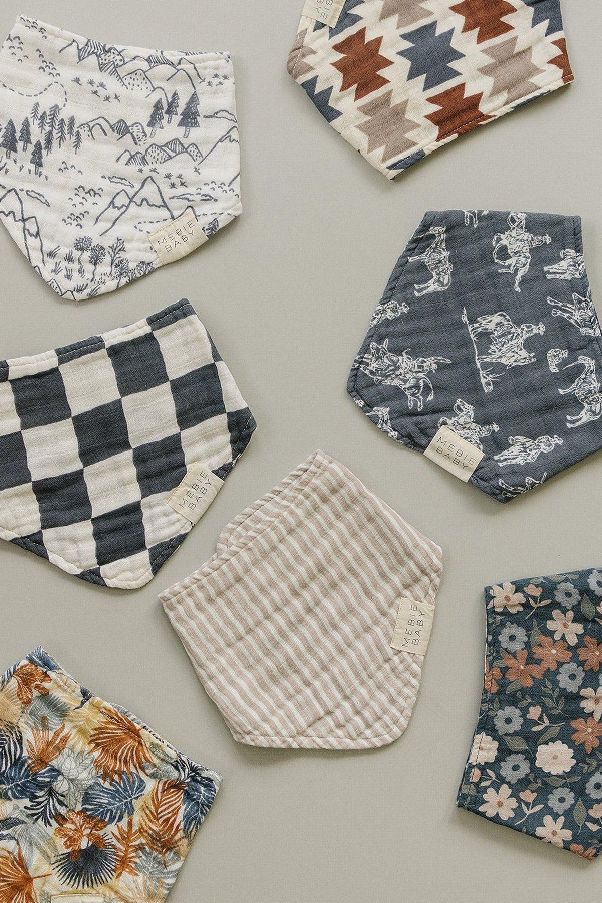 Various Mebie Baby muslin bibs in different patterns, including stripes, florals, checkered, and Aztec designs, displayed on a surface.