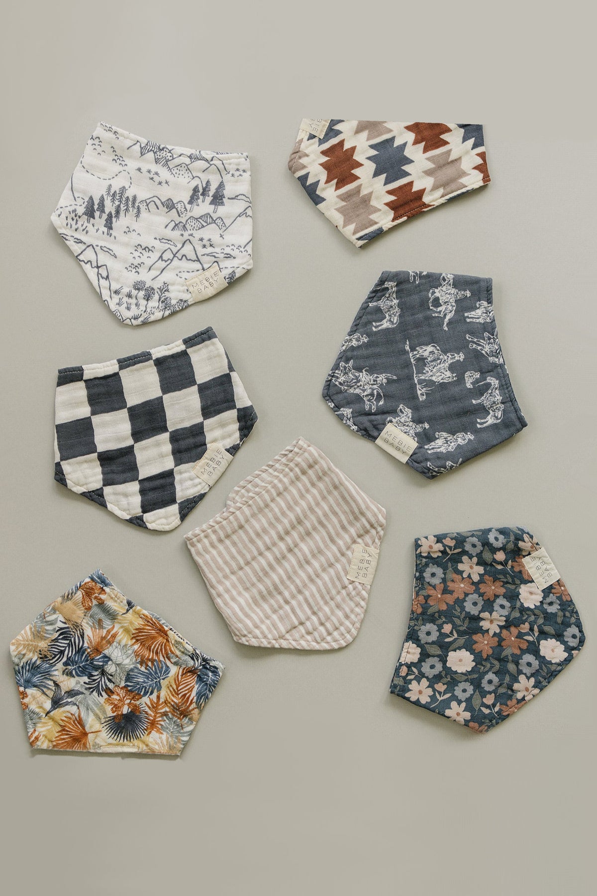 Seven Mebie Baby muslin bibs with various prints, including floral, checkered, and striped, arranged on a neutral background.