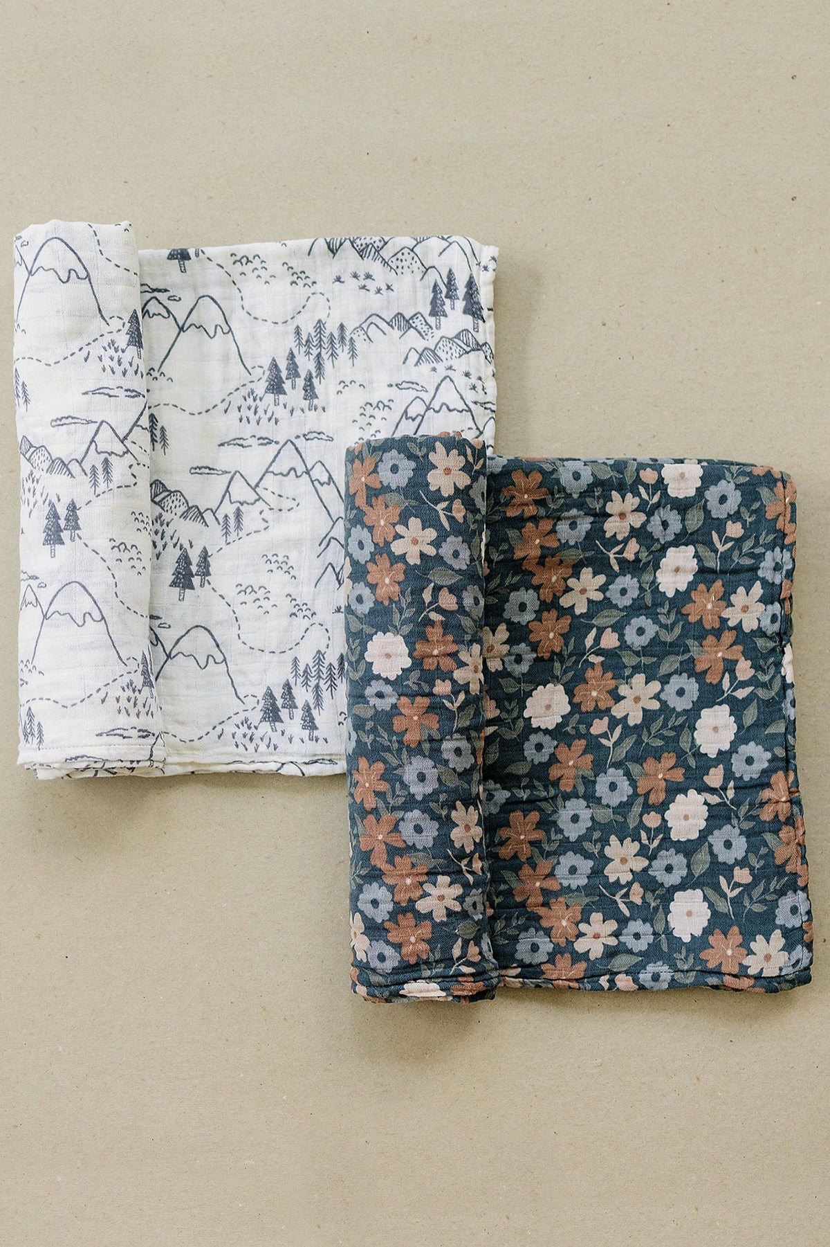 Two muslin swaddle blankets are displayed, one with a mountain pattern, the other with a floral design.
