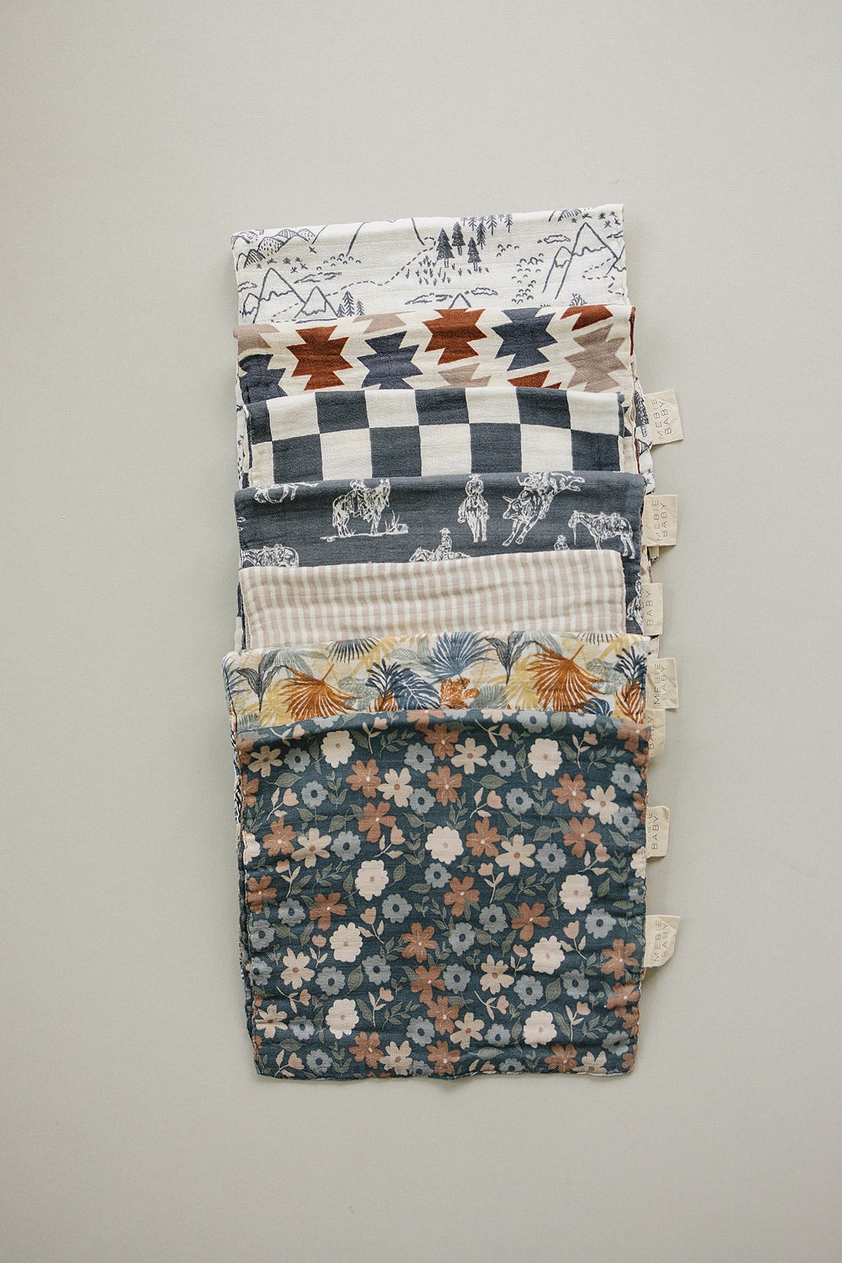 A stack of colorful muslin burp cloths featuring various patterns, including floral, geometric, and wildlife designs, displayed on a neutral.