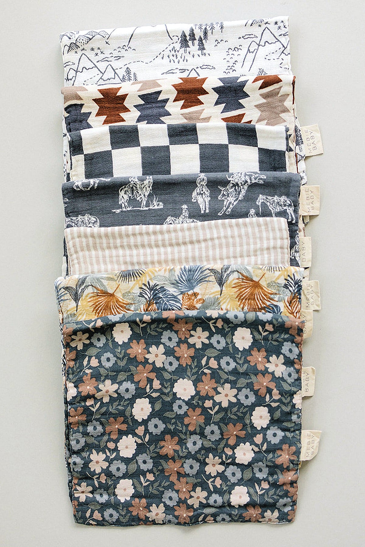 A stack of muslin burp cloths featuring various patterns, including florals, stripes, and geometric designs, in earthy colors.