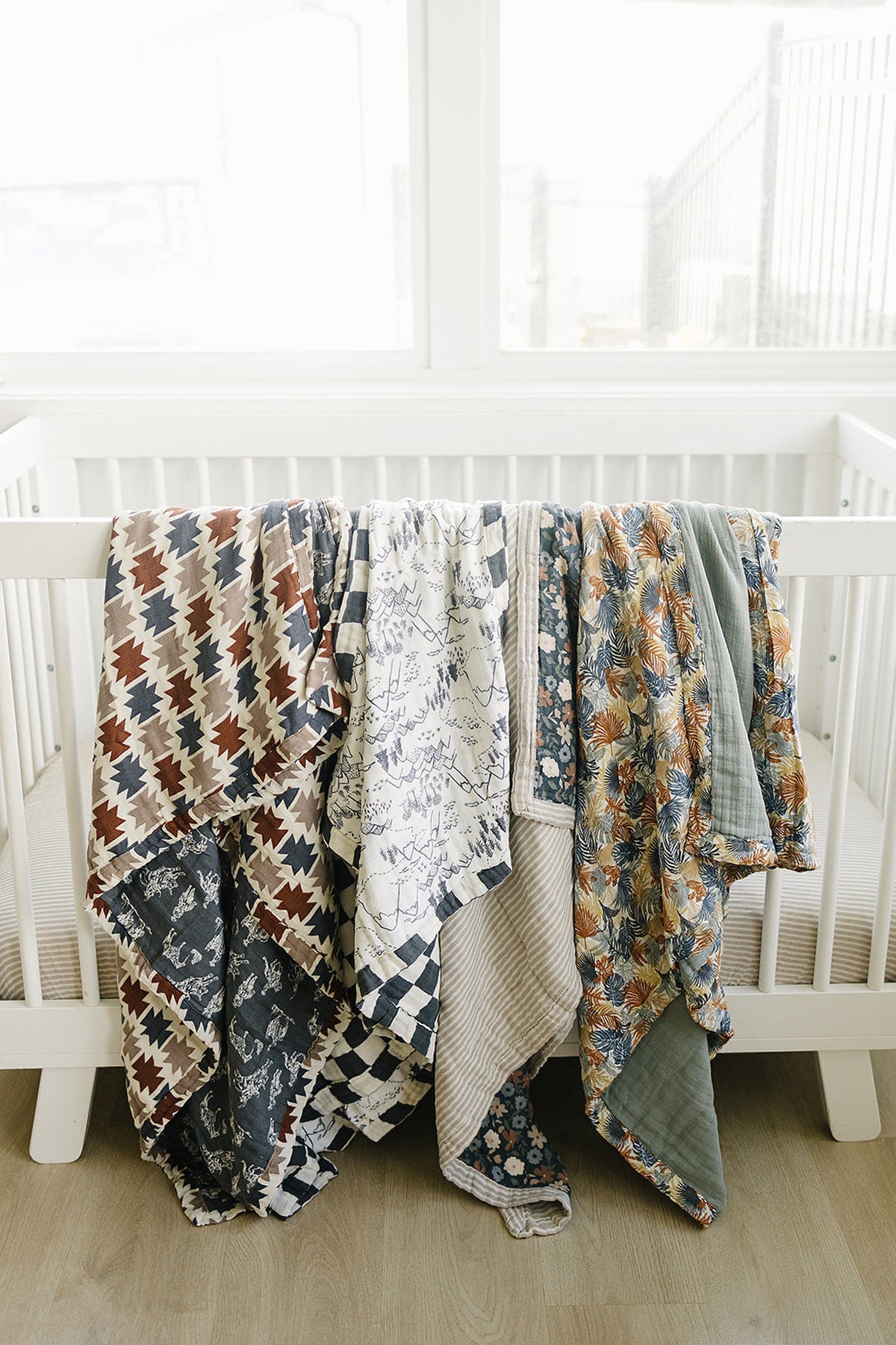 A variety of colorful muslin quilts hang over a white crib, showcasing floral and geometric patterns in soft, cozy fabrics.