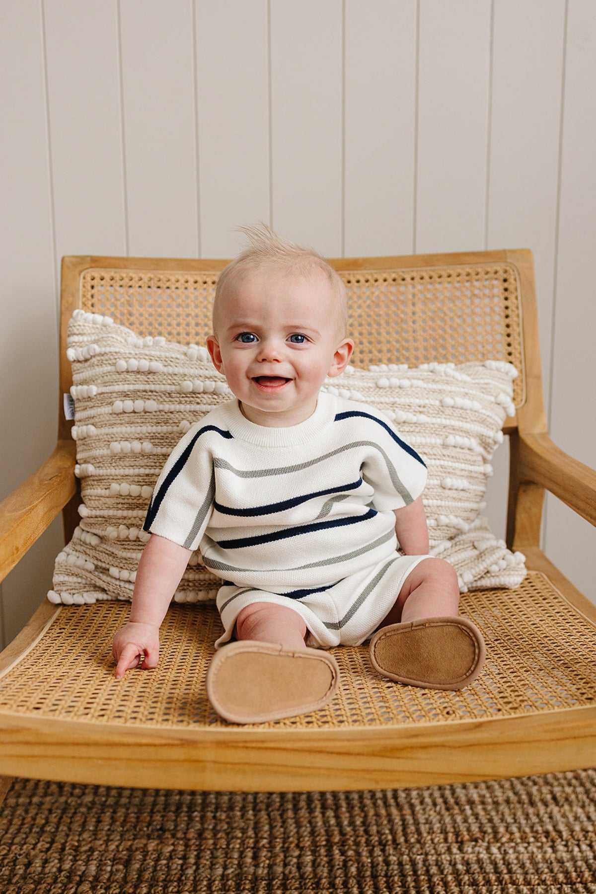 A baby wearing a striped knit outfit sits on a wooden chair with a textured cushion, smiling.