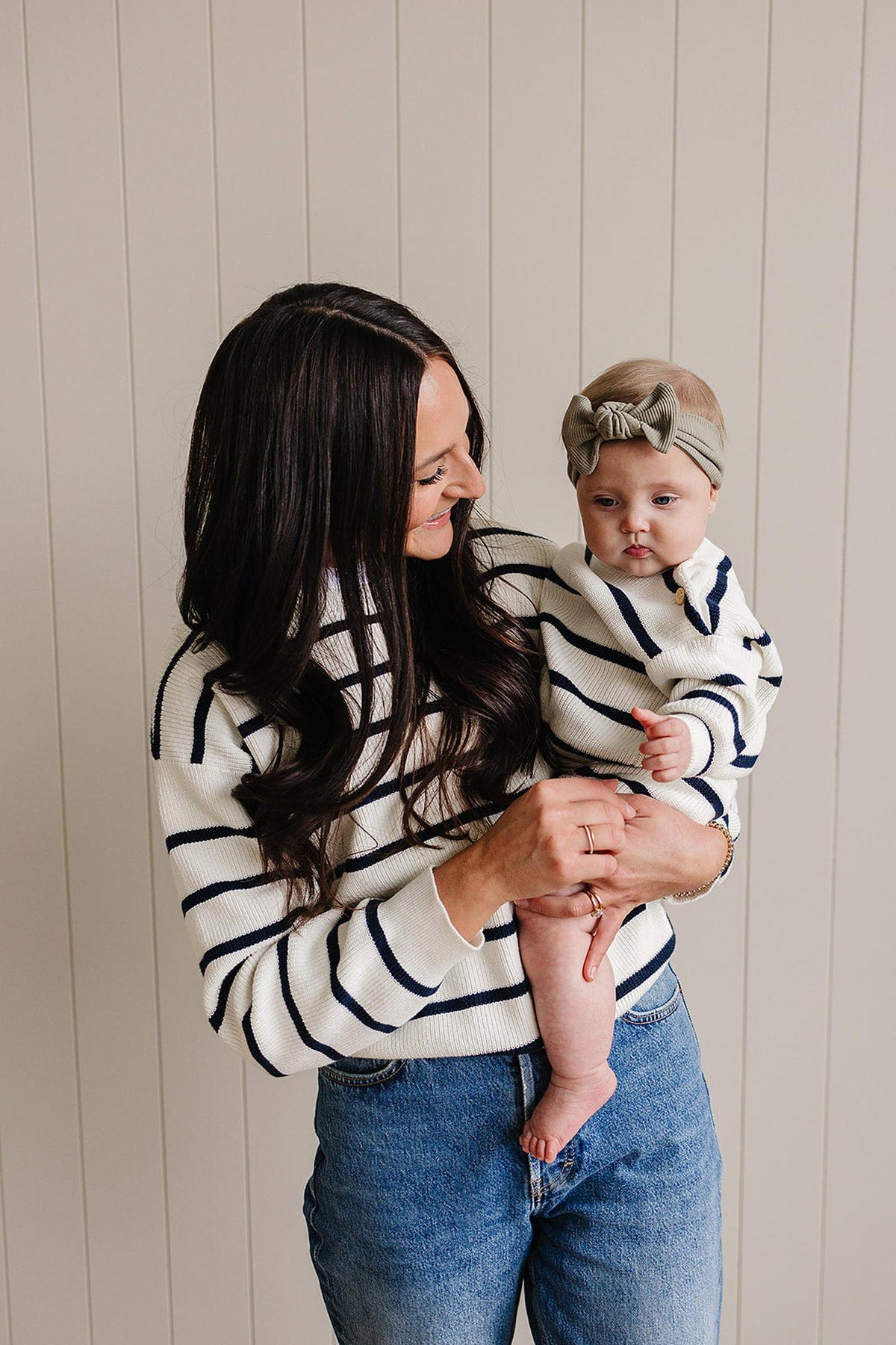 Woman holding baby, both wearing matching navy-striped sweaters, standing against a beige, paneled wall.