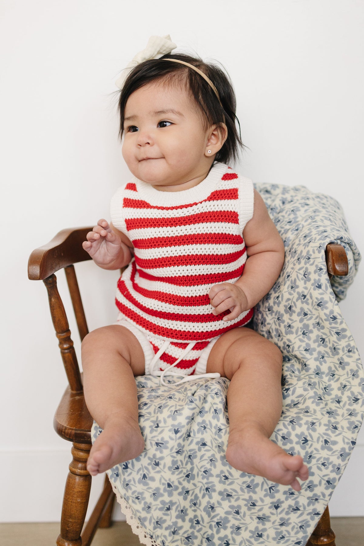 A baby girl sits on a wooden chair, wearing a red and white striped knitted bloomer tank set.