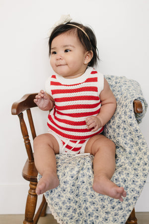 A baby girl sits on a wooden chair, wearing a red and white striped knitted bloomer tank set.