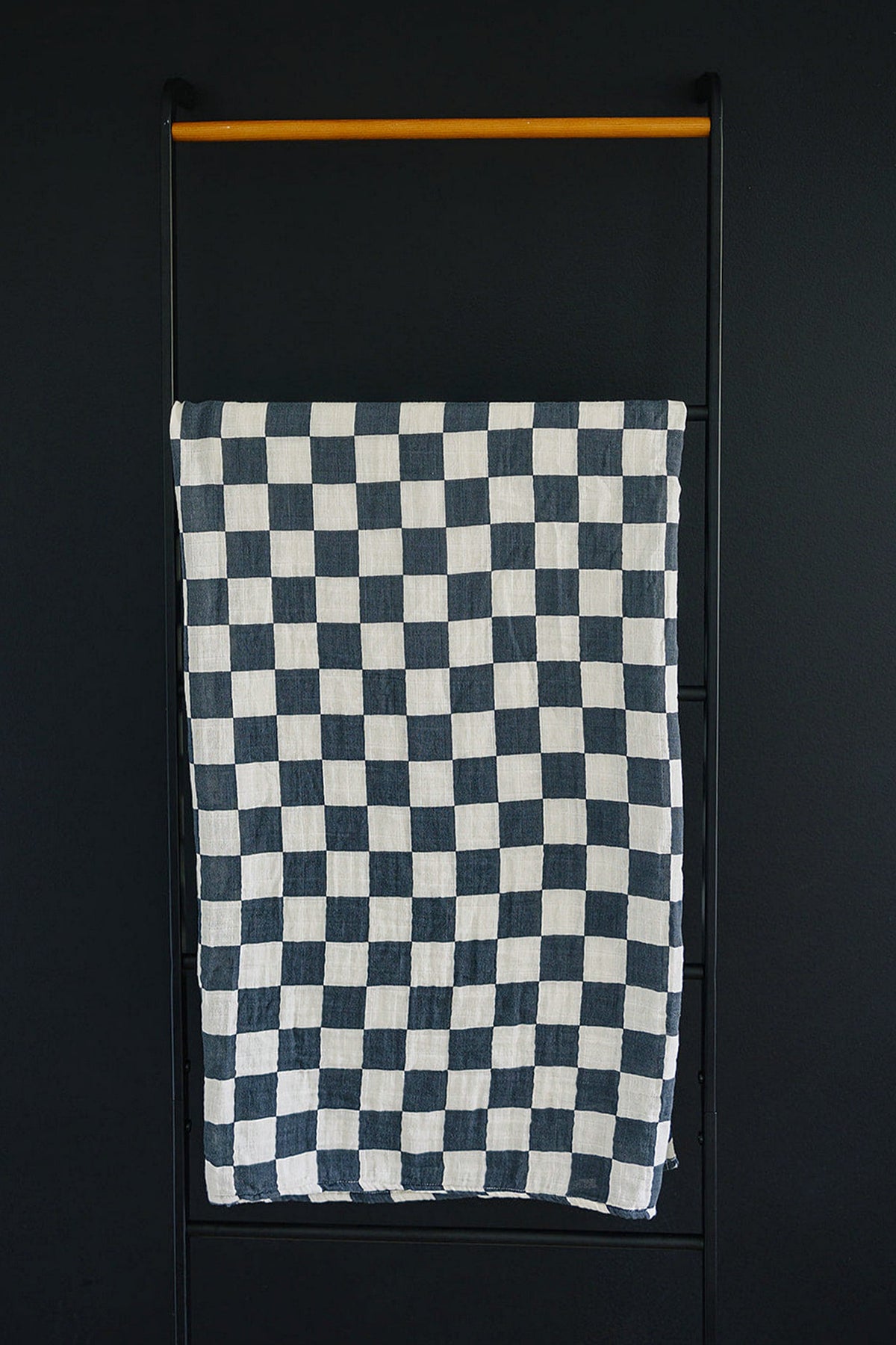 A charcoal checkered muslin blanket hangs on a black metal ladder against a dark wall.