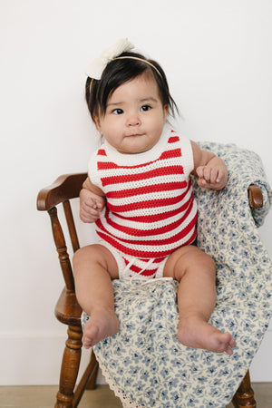 A baby girl sits on a wooden chair, wearing a red and white striped knitted tank top with a matching bloomer.