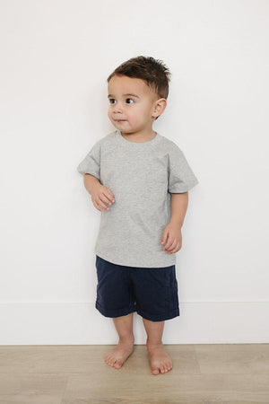 A young child stands barefoot against a white wall, wearing a grey cotton t-shirt and navy shorts.