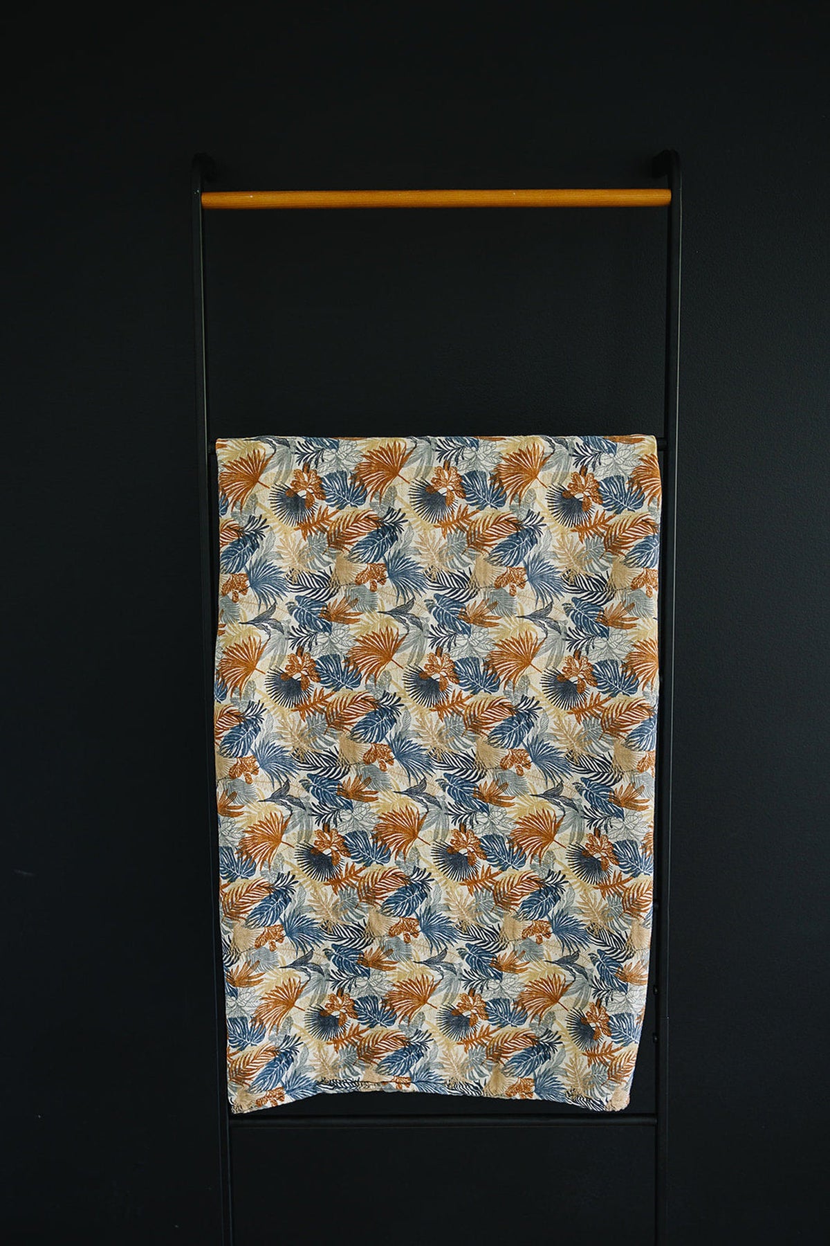 A colorful muslin swaddle blanket with a tropical leaf pattern hangs on a black and wood rack.