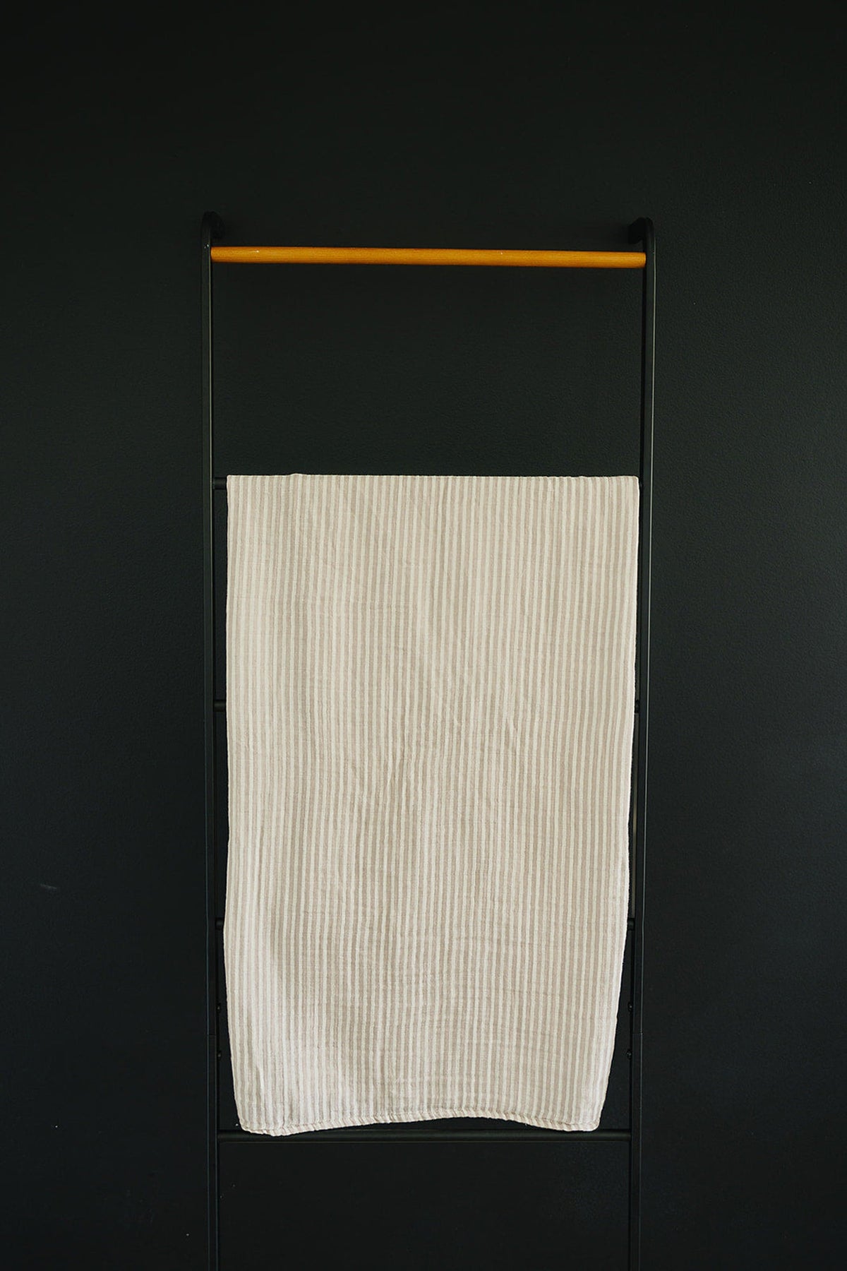 Taupe stripe muslin swaddle blanket hanging on a black metal rack with a wooden bar, against a dark background.