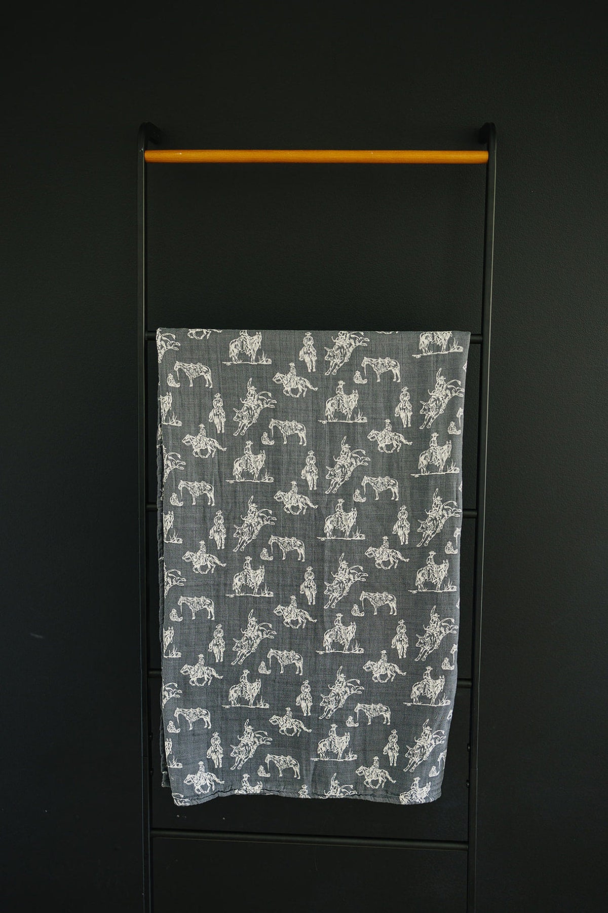 Gray muslin swaddle blanket with white Wild West-themed prints, hanging on a black metal and wood rack.