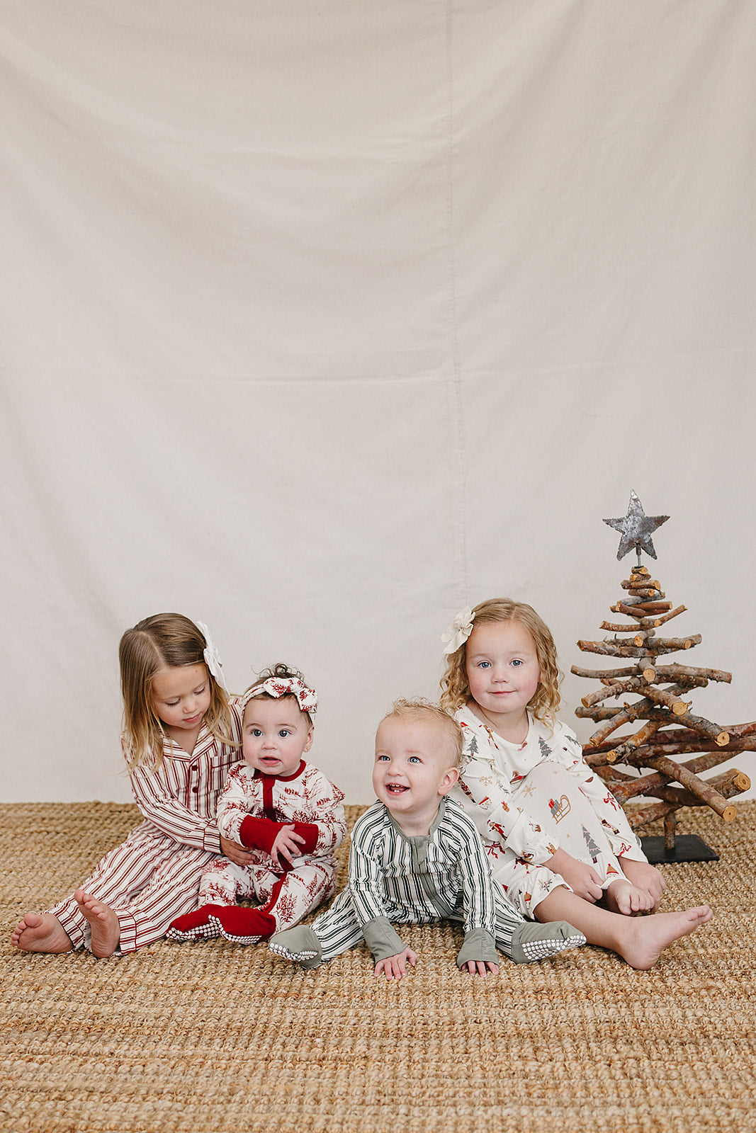 Four young children sit on a woven rug against a neutral backdrop, wearing festive pajamas in red and white stripes, green stripes, and holiday patterns, surrounded by a rustic wooden.