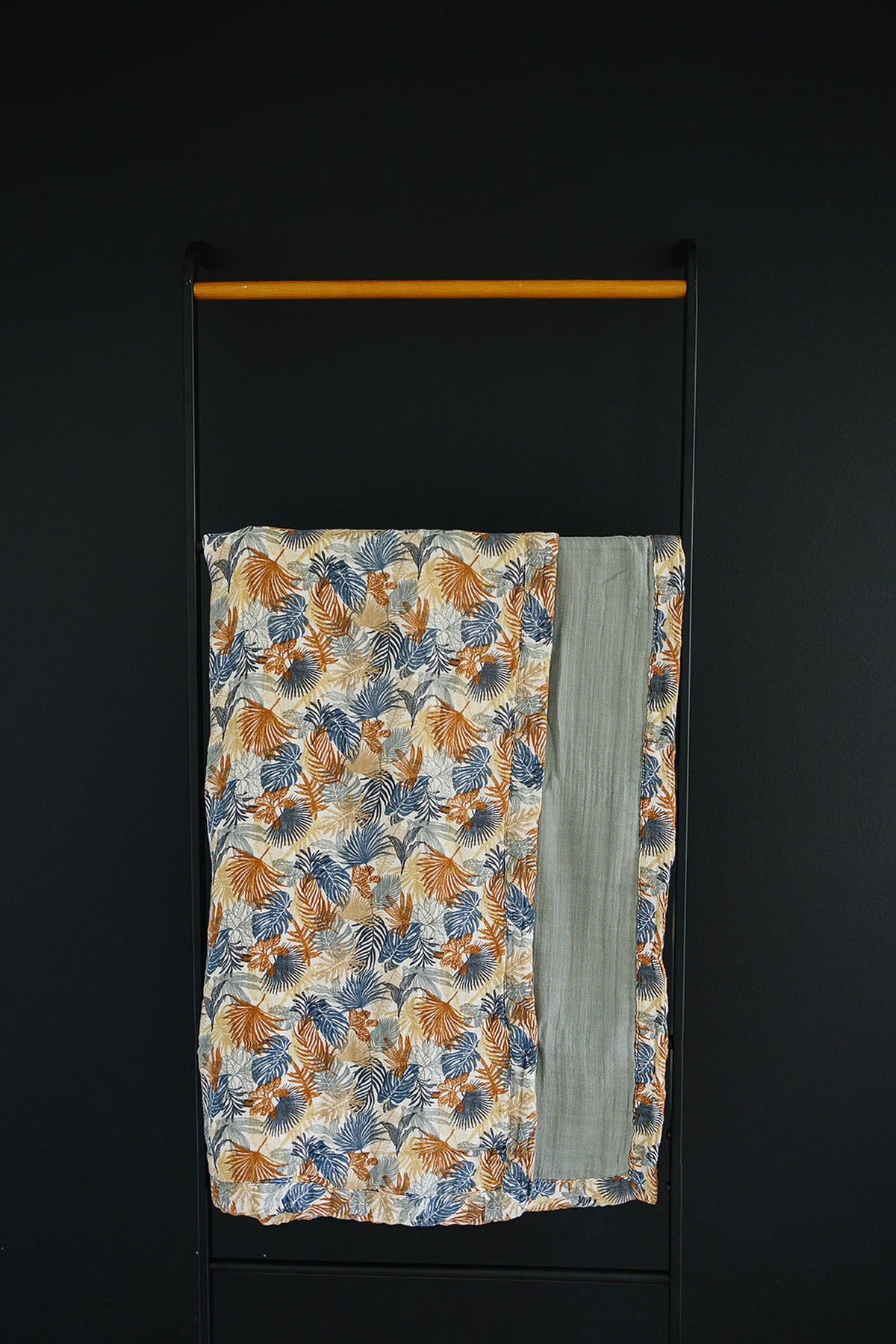 Floral-patterned muslin quilt with orange and blue palm leaves, draped over a black metal rack against a dark background.