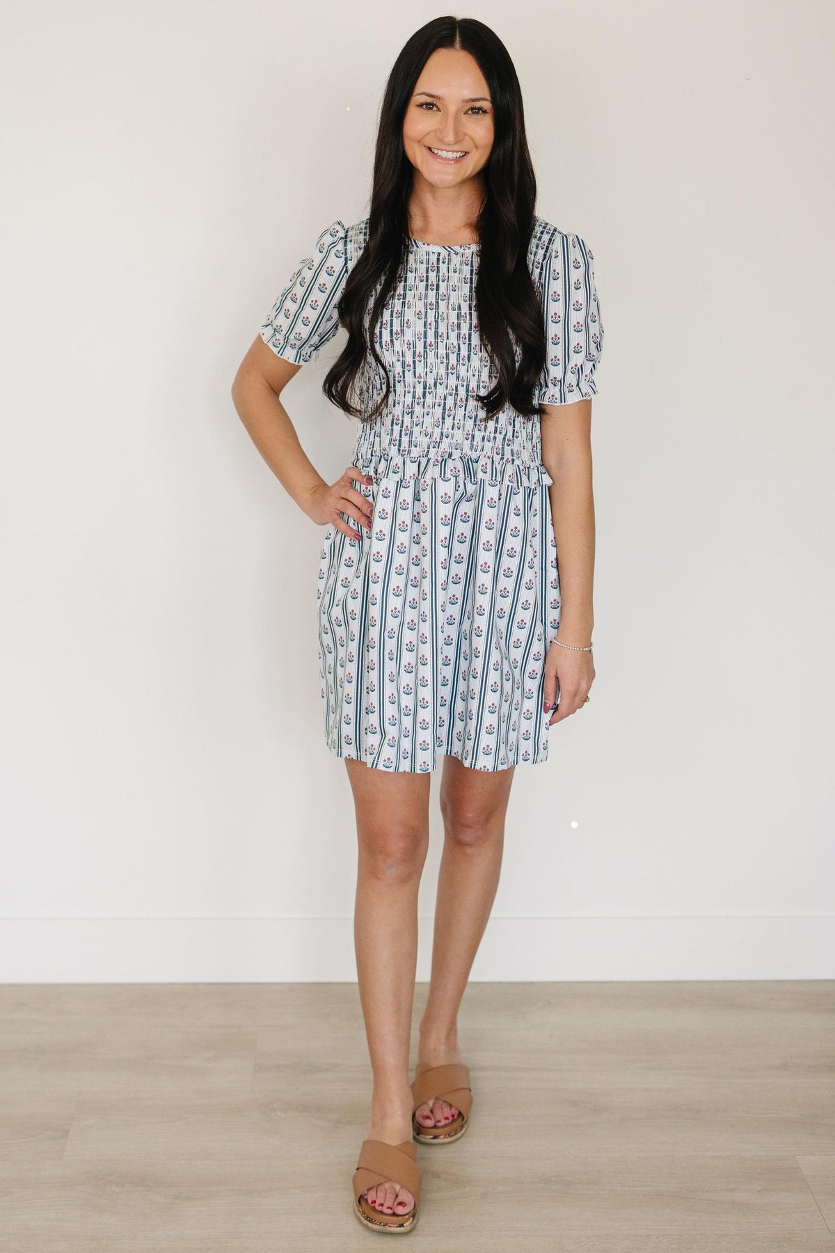 A woman stands in a light blue floral dress with a smocked bodice and puffed sleeves, showcasing a delicate pattern against a neutral background.