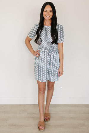 A woman stands in a light blue floral dress with a smocked bodice and puffed sleeves, showcasing a delicate pattern against a neutral background.
