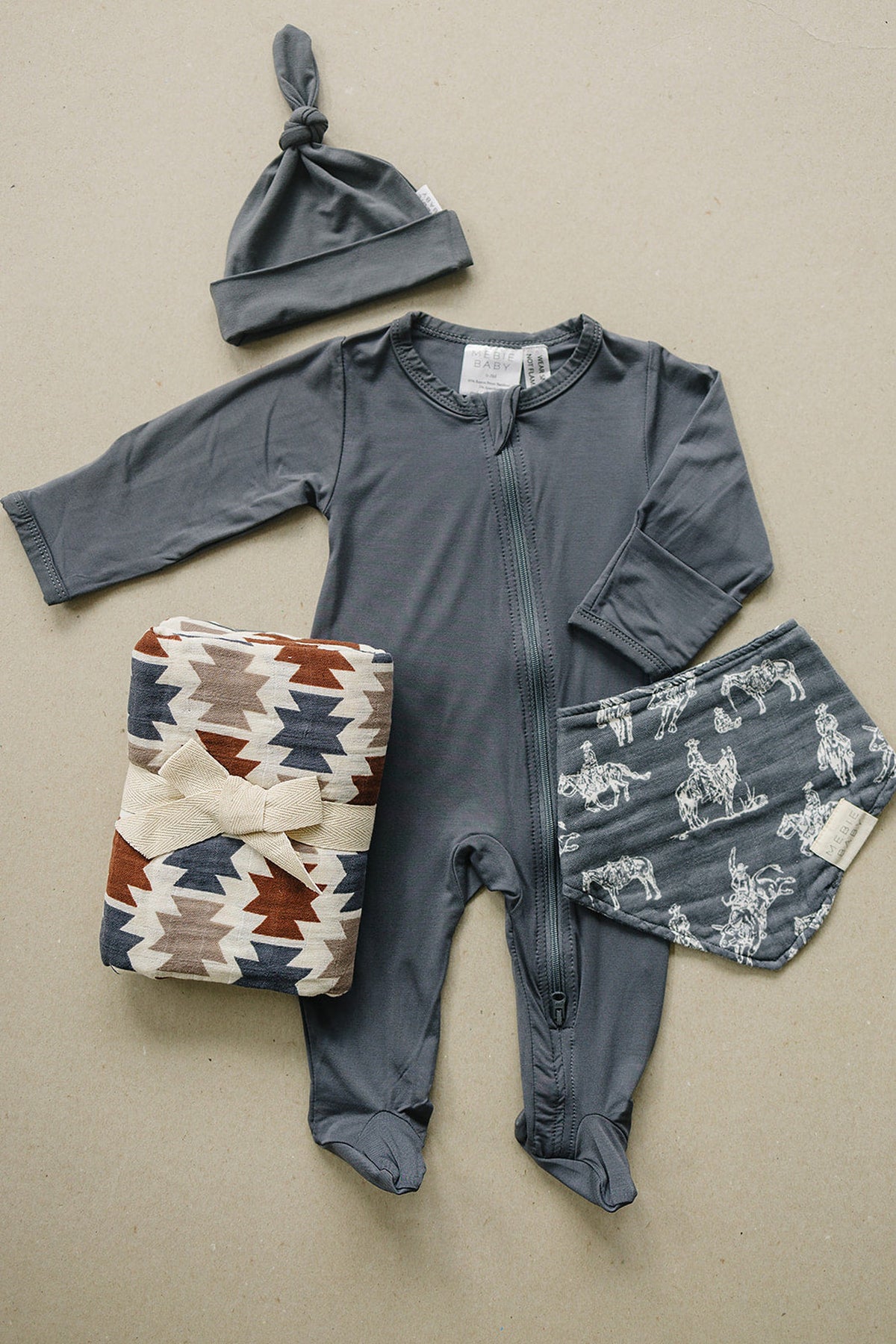 A gray zip-up onesie, a matching knotted hat, and patterned blankets are displayed on a neutral background.