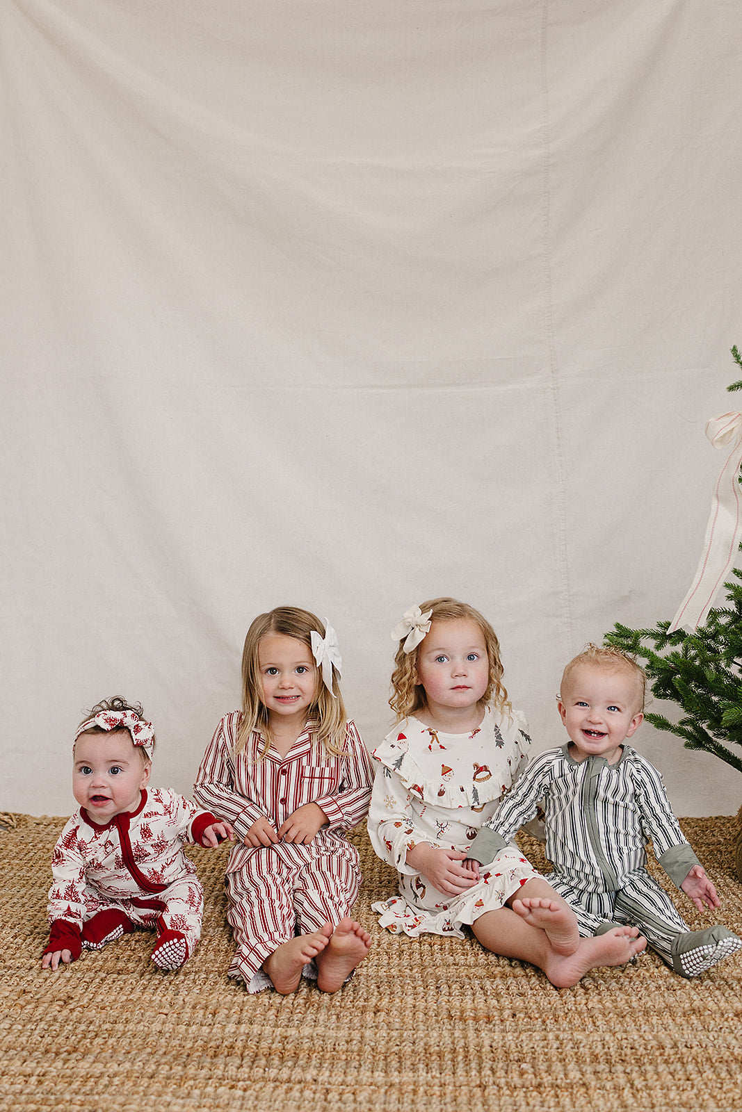 Four children sit on a natural woven mat in festive pajamas, featuring red stripes and holiday patterns, against a soft beige backdrop with a Christmas tree nearby.