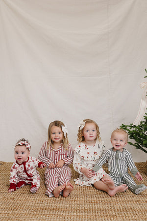 Four children sit on a woven rug in festive holiday gowns, featuring red and white stripes, a floral pattern, and green stripes.