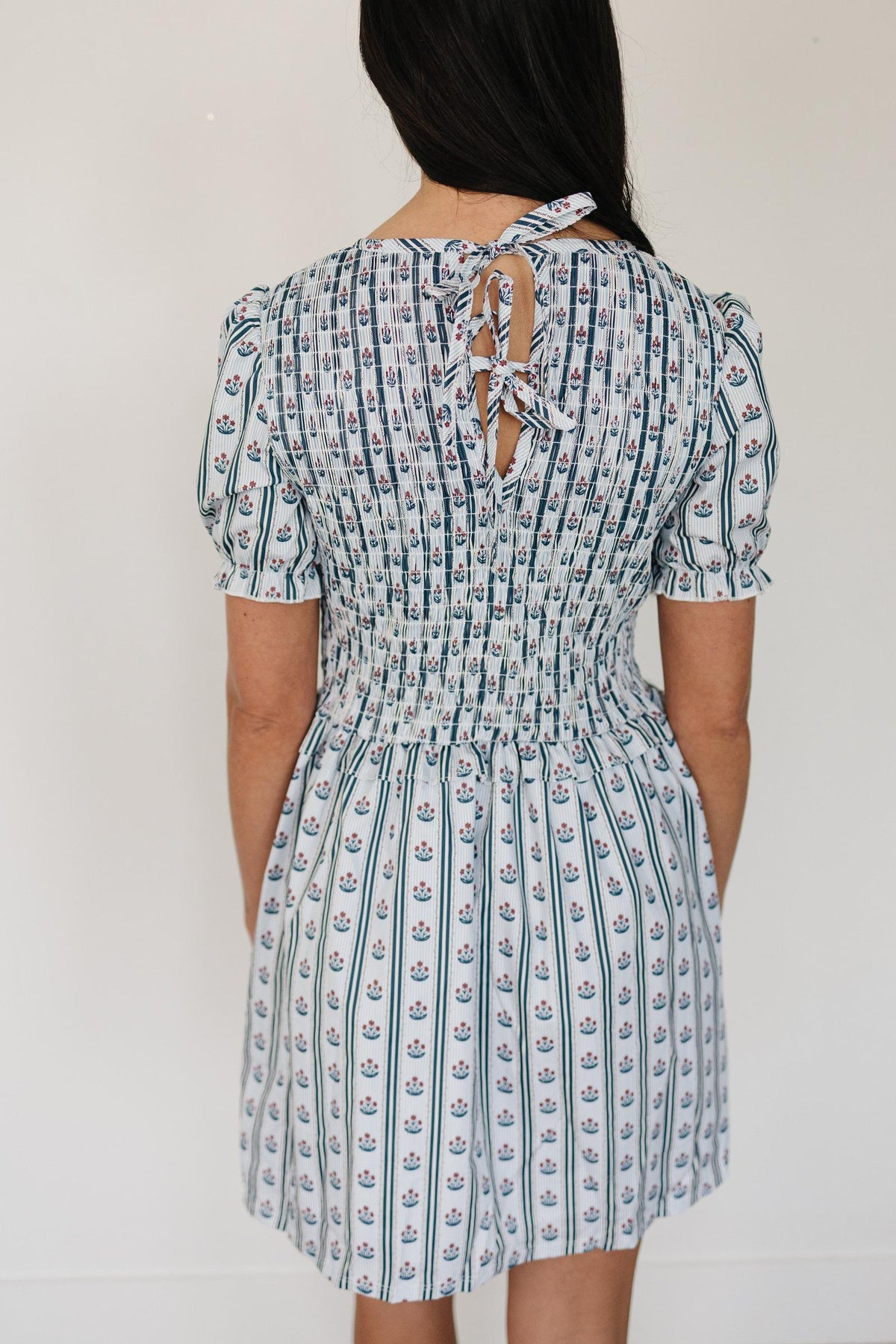 A woman is shown from the back wearing a short-sleeved, floral block print dress with a smocked waist and a tie closure at the neck.