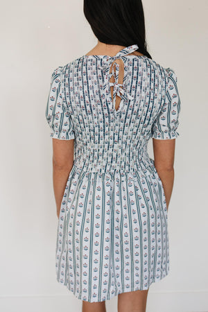 A woman is shown from the back wearing a short-sleeved, floral block print dress with a smocked waist and a tie closure at the neck.