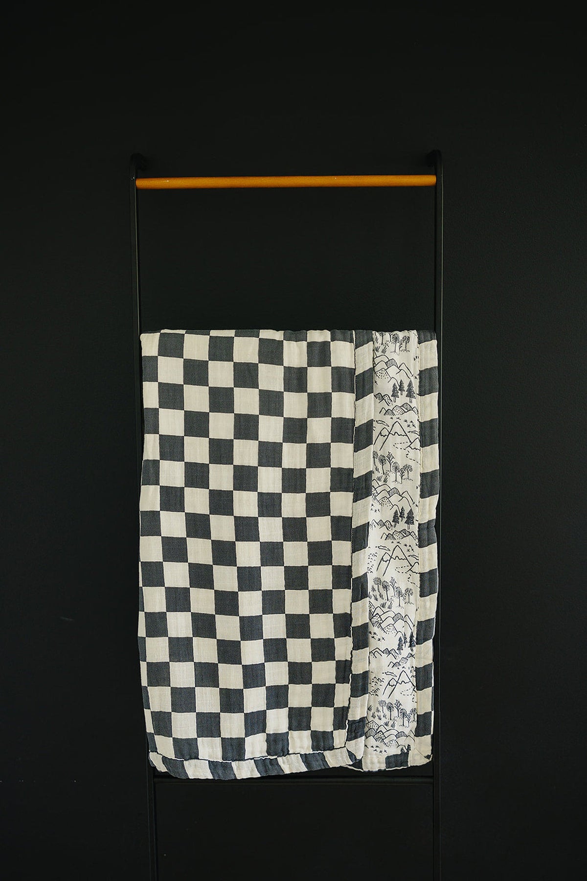 A charcoal checkered muslin quilt hangs on a black rack, featuring a detailed landscape print on one edge.