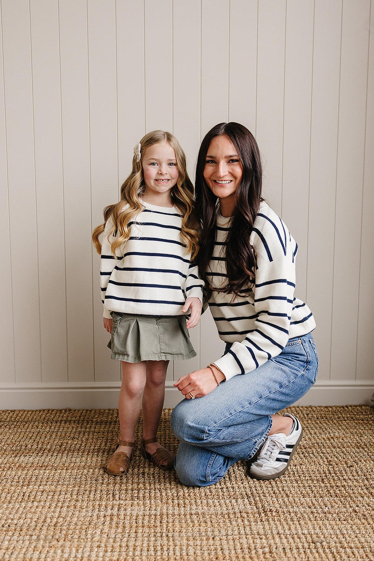 A woman and child wearing matching navy striped sweaters pose on a woven rug against a beige paneled wall.