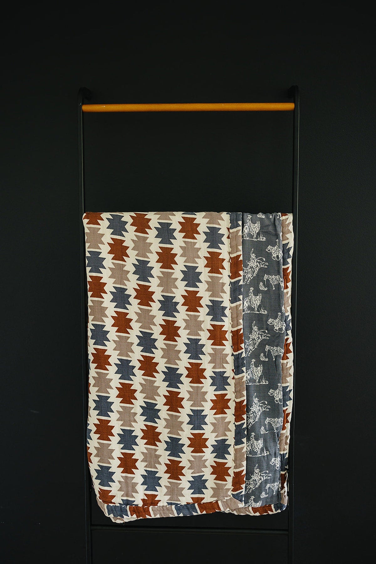 Aztec-patterned muslin crib quilt in earthy tones, featuring a cowboy-themed border, hanging on a minimalist black rack.