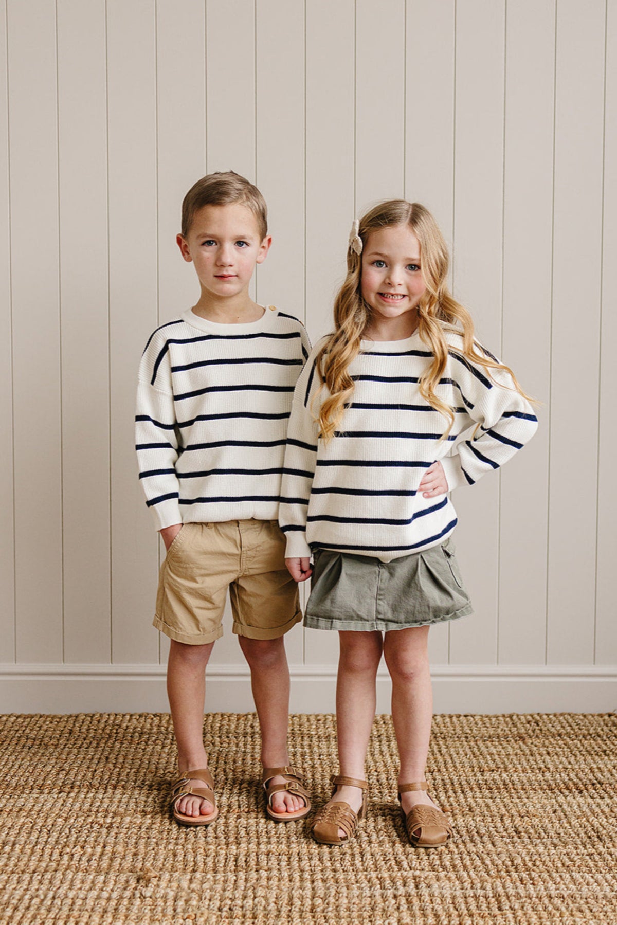Two children in navy striped sweaters, one wearing khaki shorts, the other a green skirt, standing indoors.
