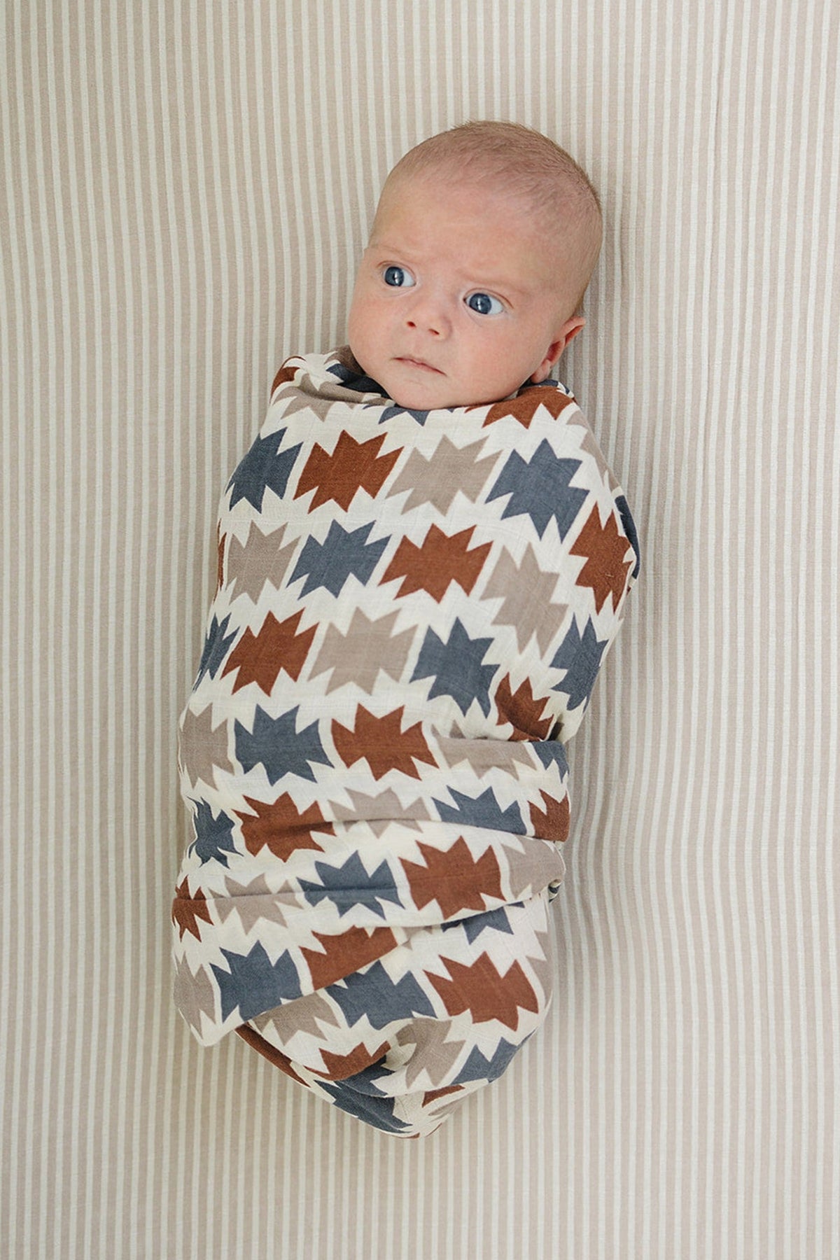 A swaddled baby with blue eyes is wrapped in a Western Aztec muslin blanket featuring earthy tones against a striped.