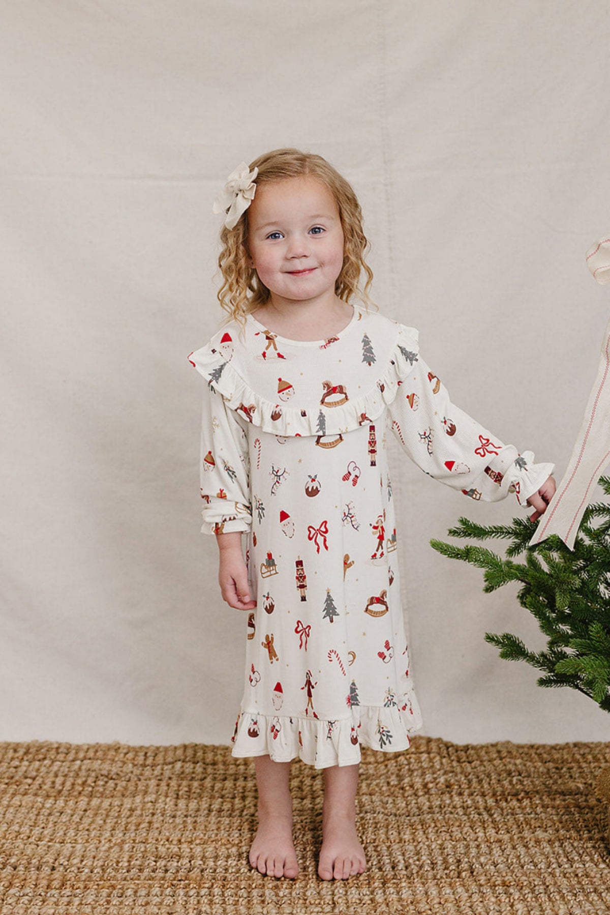 A young girl stands in a festive, cream-colored bamboo gown adorned with holiday motifs. She holds a ribbon next to a small Christmas tree, showcasing a cozy and cheerful holiday.