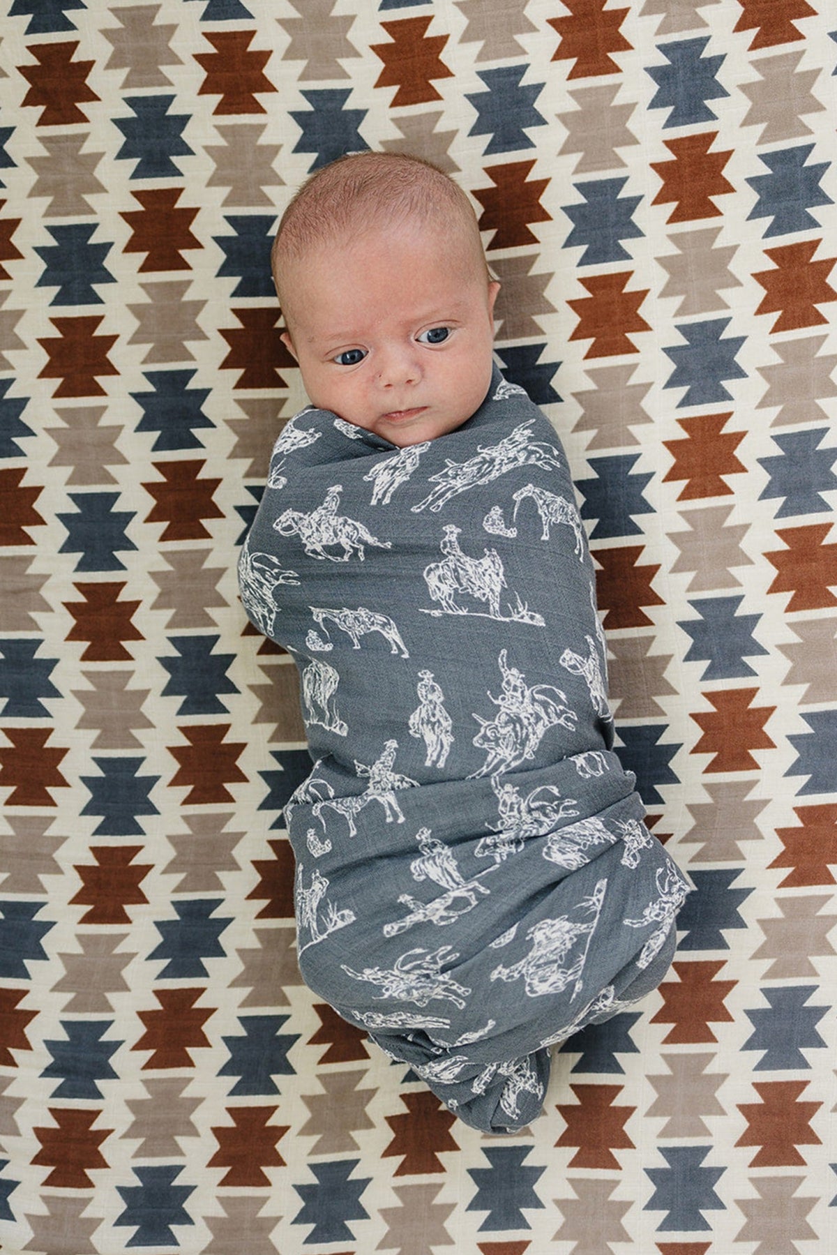 A baby is swaddled in a gray muslin blanket with Western prints, lying on a patterned textile.
