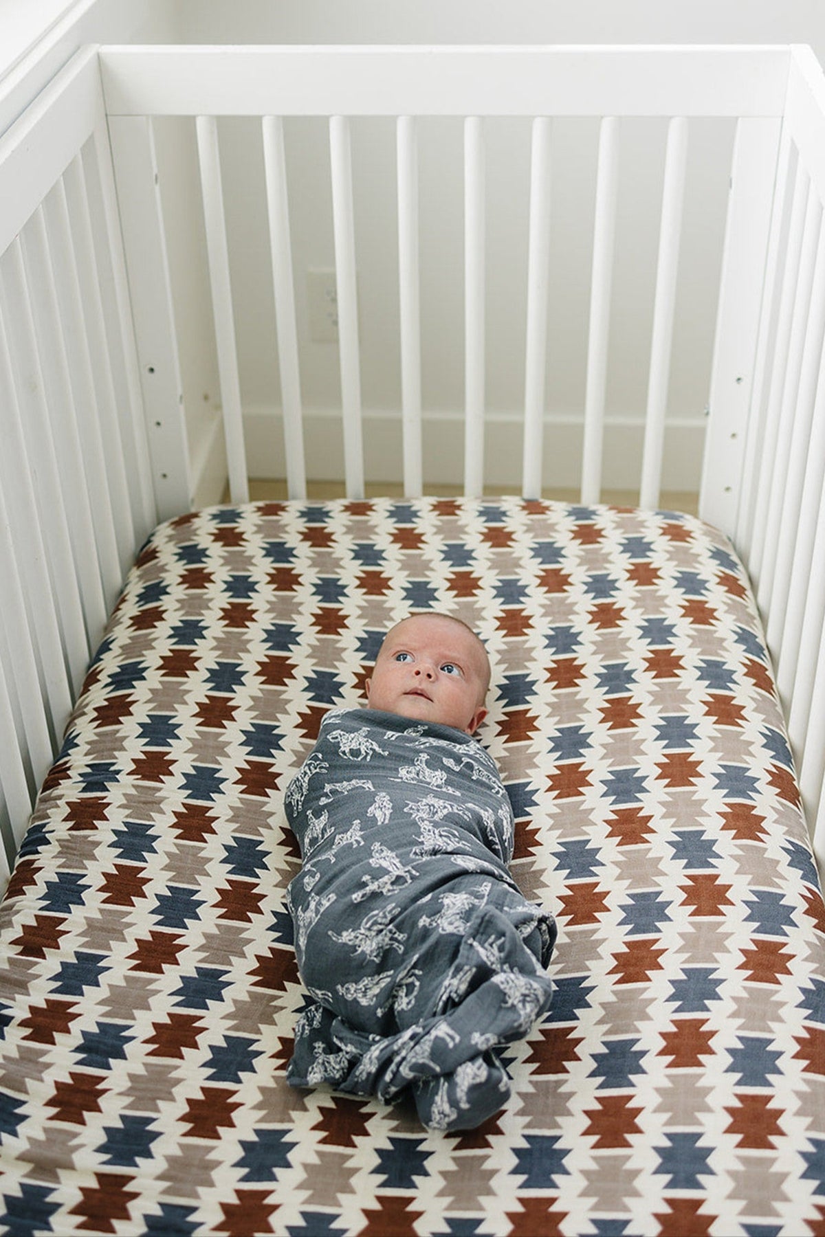 A wrapped baby lies on a colorful Western Aztec patterned crib sheet in a white crib, surrounded by soft, breathable.