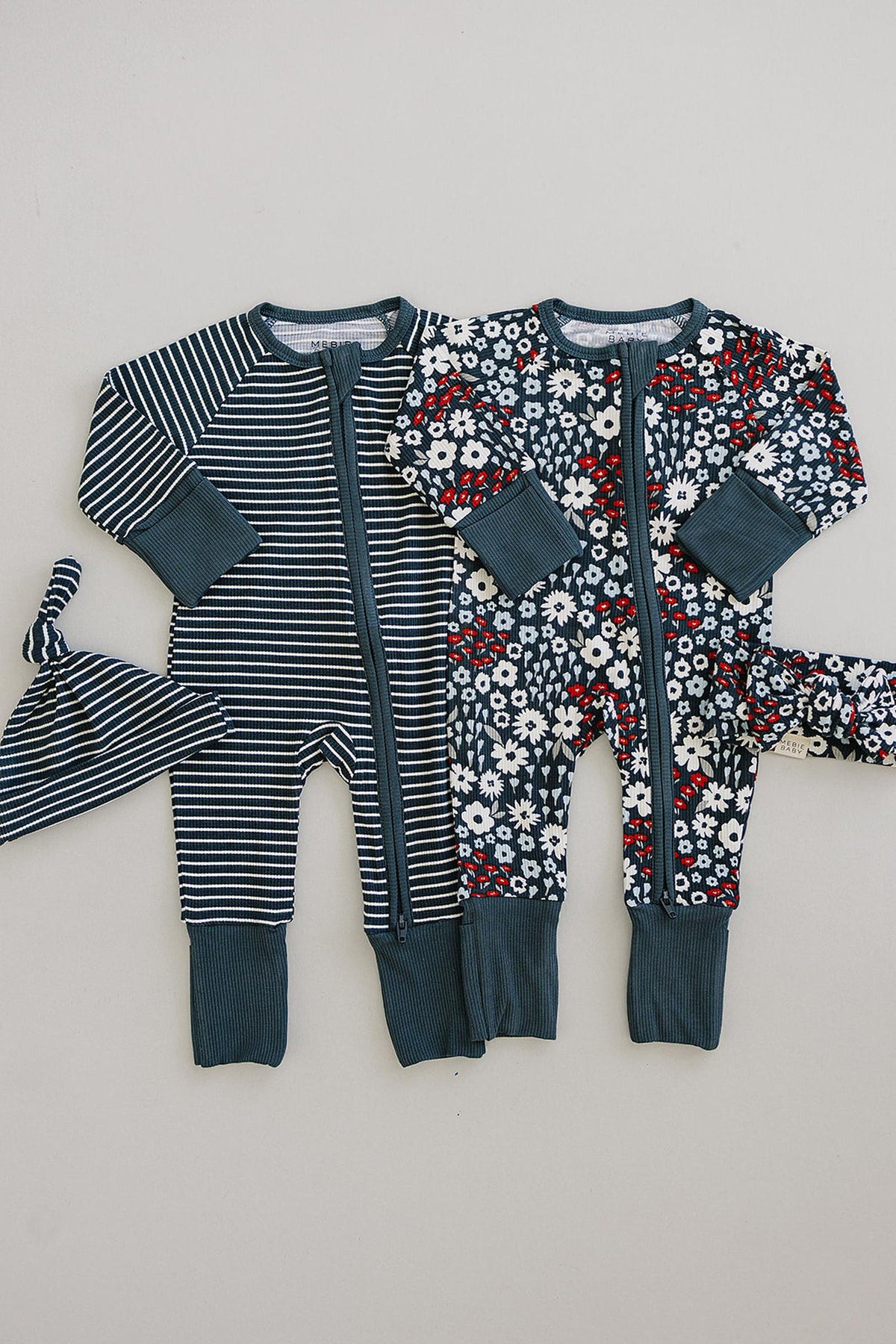 Two baby pajamas with navy stripes and floral patterns, featuring ribbed cuffs and a matching knotted hat.