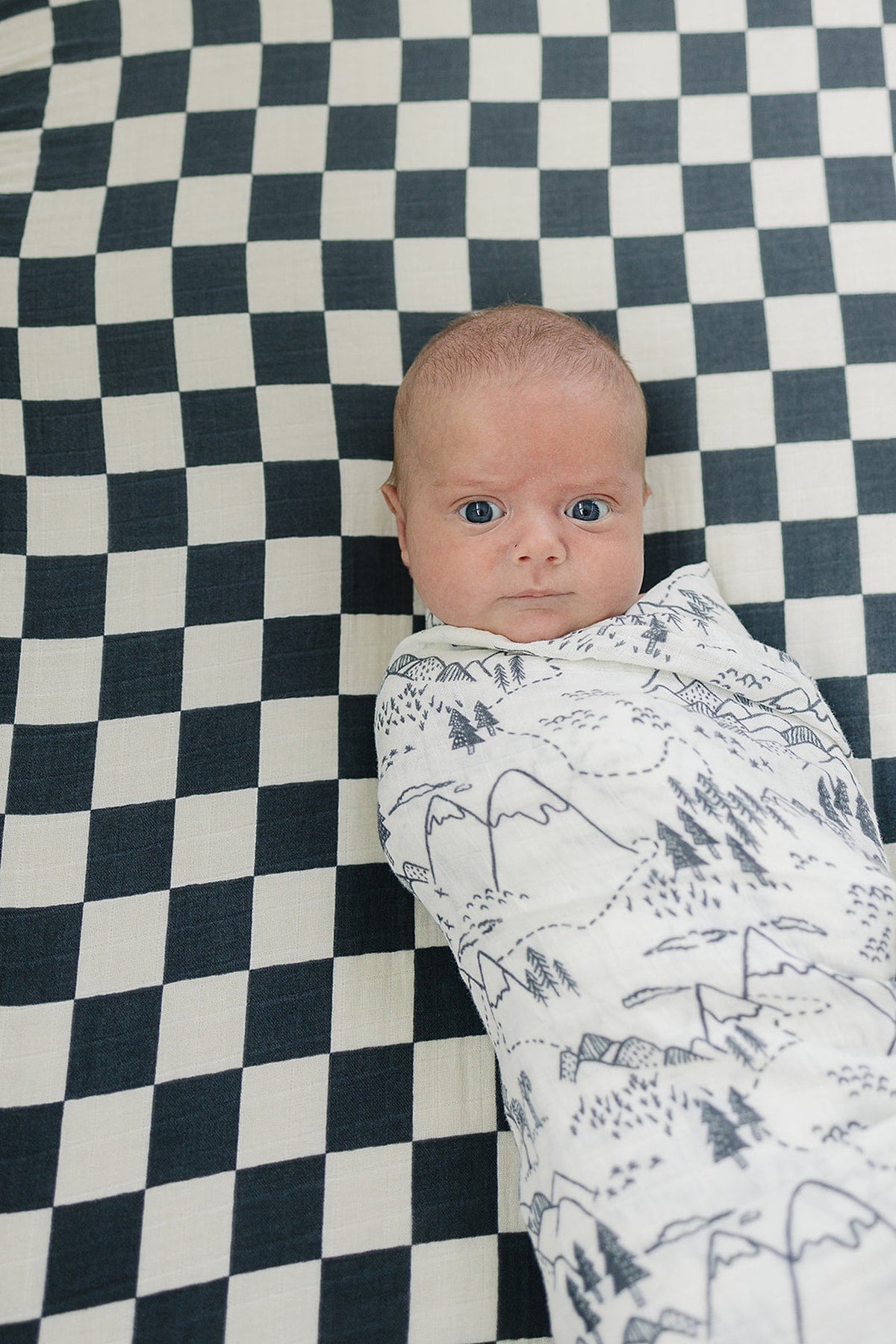 A baby wrapped in a white muslin swaddle with mountain designs, lying on a black and white checkered blanket.