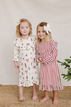 Two young girls stand together, one wearing a white gown with festive prints and the other in a red and white striped dress.